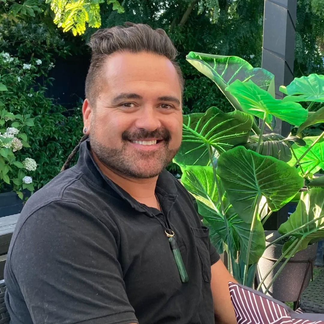 A smiling man with short brown hair, beard, and earrings, sitting outdoors next to large green and tropical plants.