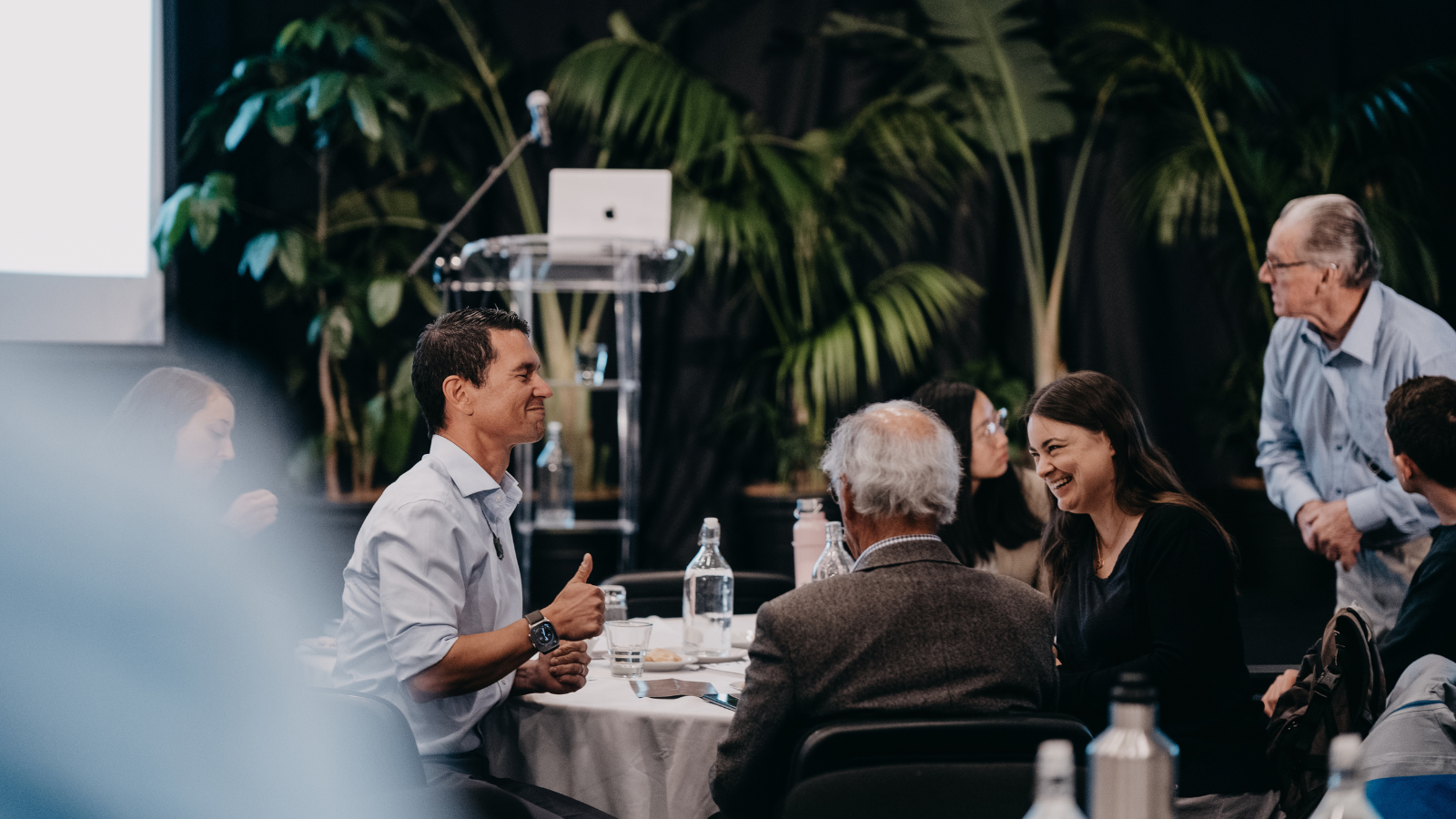 People sitting around a table in a conference room with plants in the background, participating in a discussion, one man smiling and giving a thumbs-up, others smiling and listening.