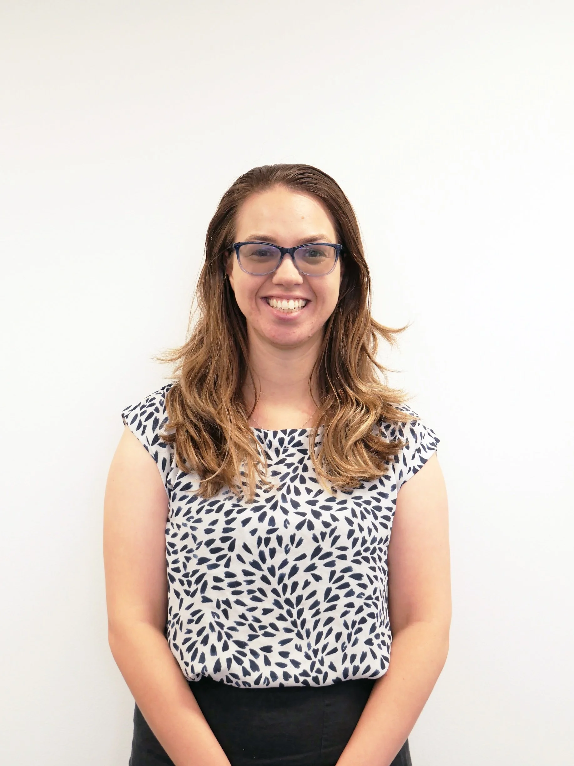 A photo of Emma Brouwer with long wavy brown hair, wearing glasses and a black-and-white patterned short-sleeve blouse, smiling and standing against a plain white wall.