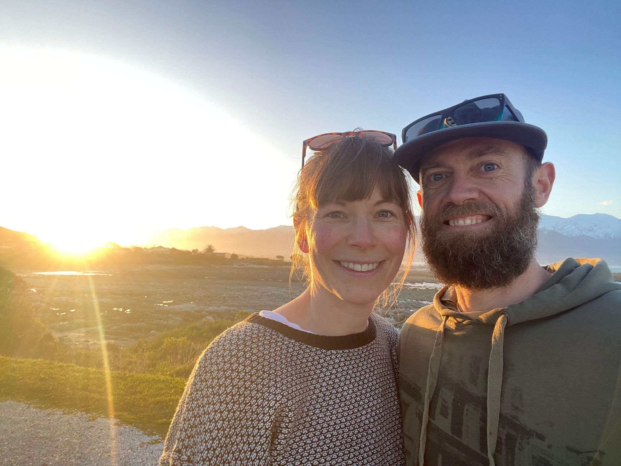 A smiling couple taking a selfie outdoors during sunset, with mountains and a rural landscape in the background. The woman has red hair and is wearing sunglasses on her head, while the man has a beard and is wearing a cap.