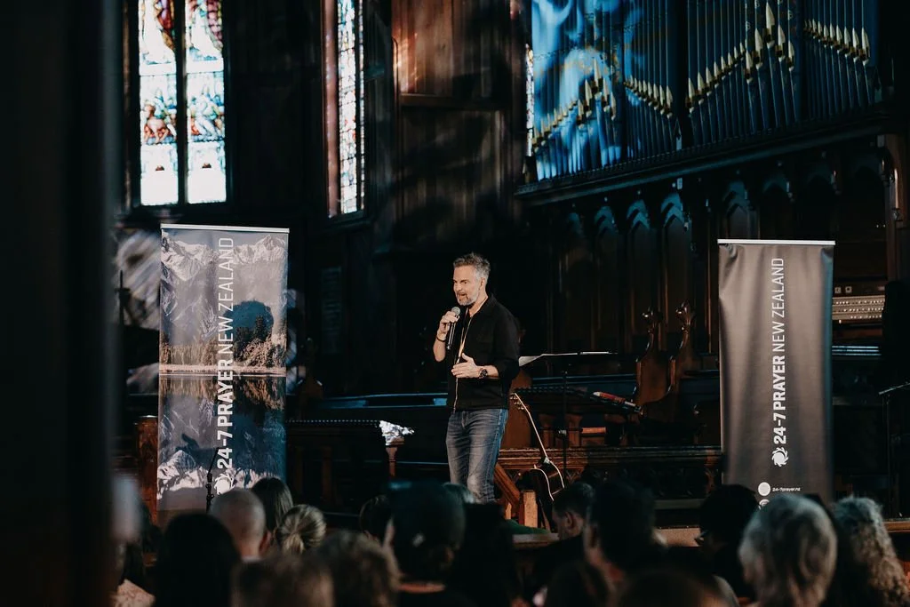 A man speaking into a microphone on a stage inside a church with stained glass windows and wooden architecture, with an audience seated in front. There are banners on either side of the stage that read '24-7 PRAYER NEW ZEALAND'.