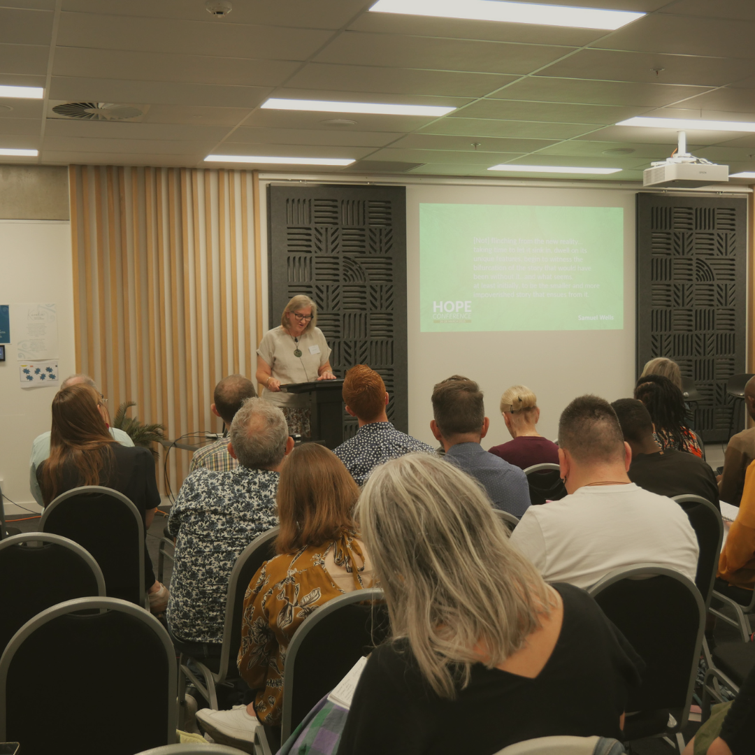 A woman giving a presentation in a conference room with an audience seated in chairs, a large screen displaying a slide, and black acoustic panels on the wall.