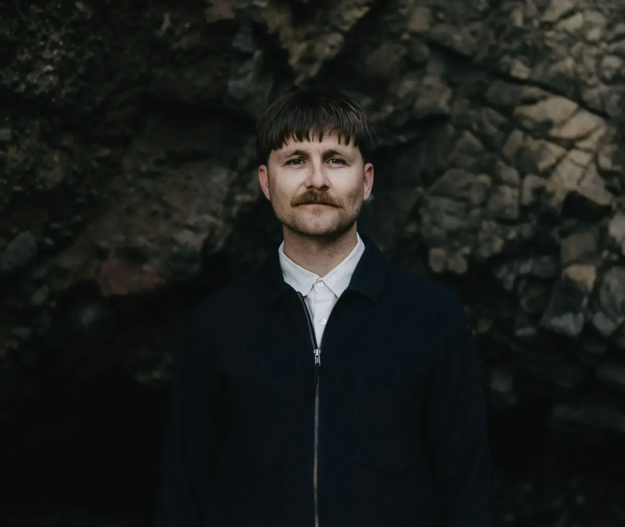 A man with dark hair and a mustache, wearing a dark jacket and white shirt, standing near rocky terrain.