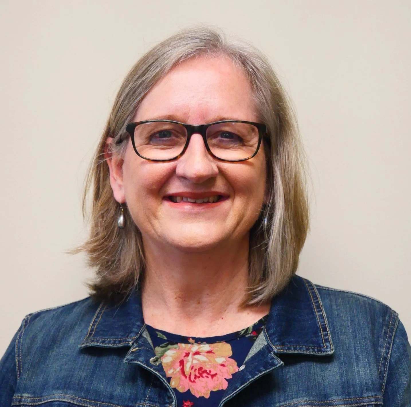 A woman with shoulder-length gray hair, wearing glasses, earrings, a denim jacket, and a floral top, smiling against a plain beige background.