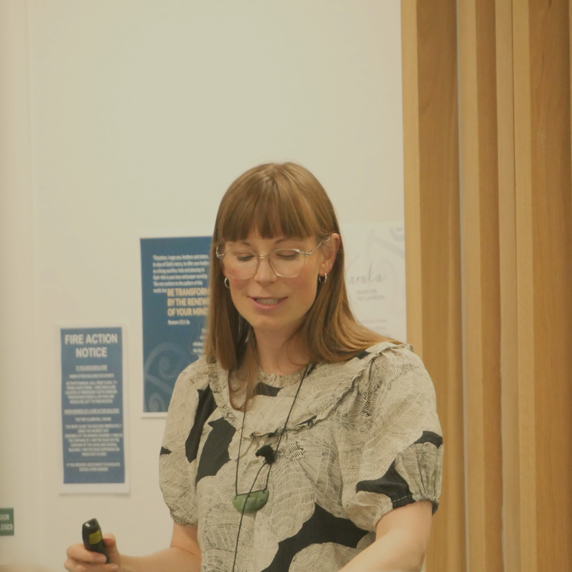 A woman with glasses and earrings standing in a room, holding a remote, with posters on the wall behind her.