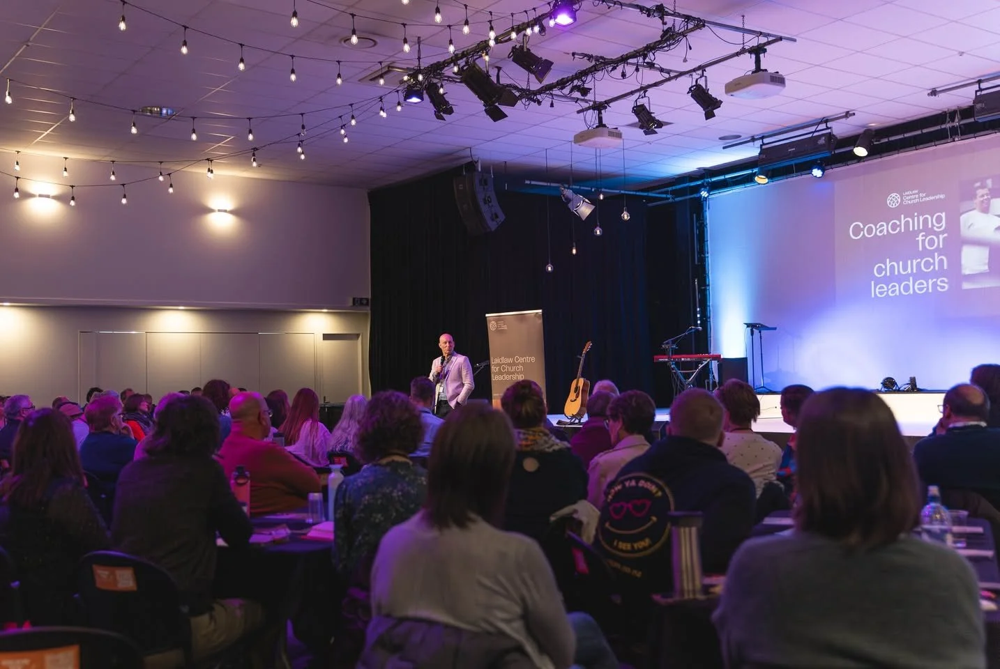 People sitting in an auditorium attending a presentation on coaching for church leaders. A speaker stands on stage with a guitar beside him, and a projection displays the title of the session.