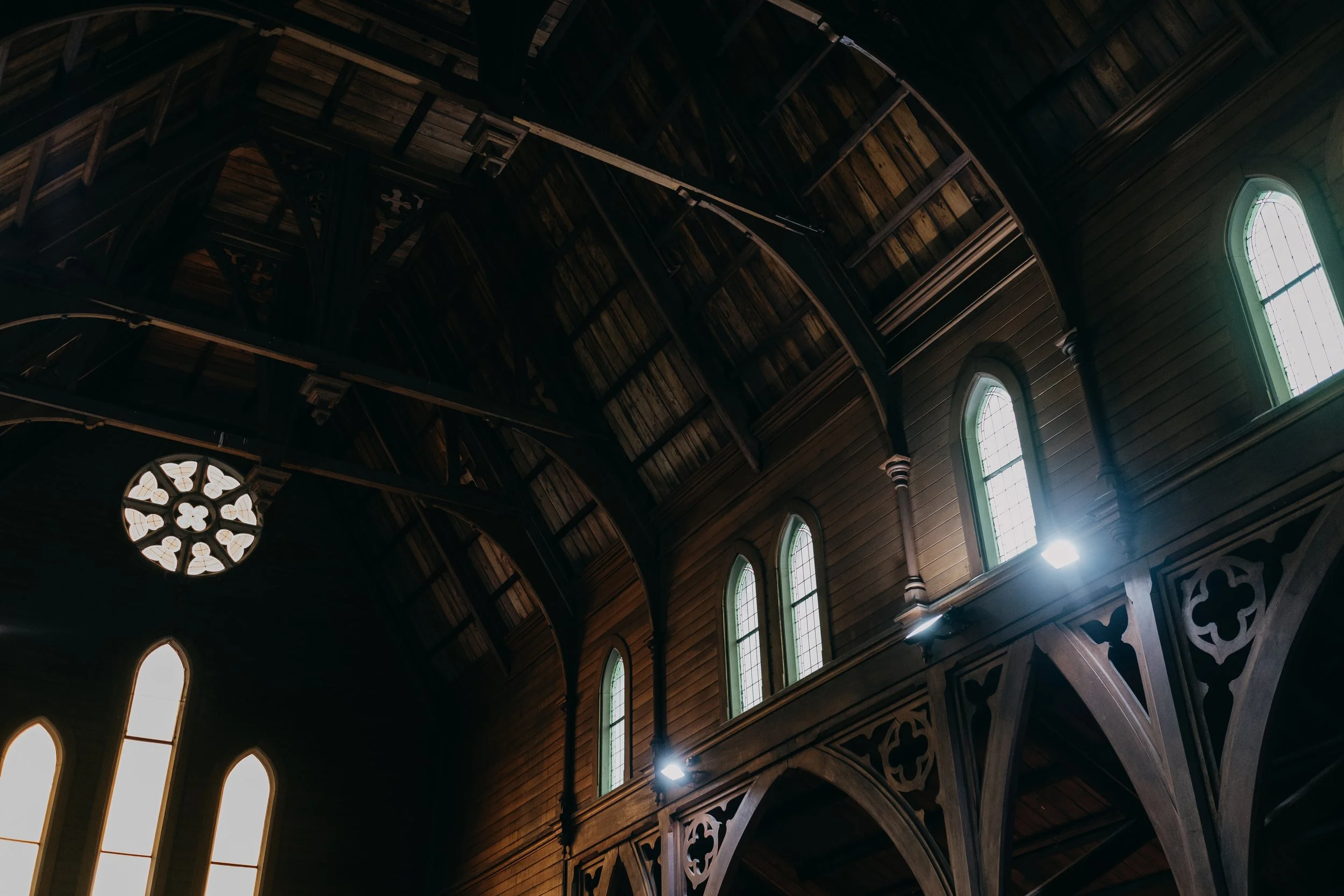 Interior view of a church ceiling with wooden beams, tall arched windows with stained glass, and decorative woodwork on the walls.