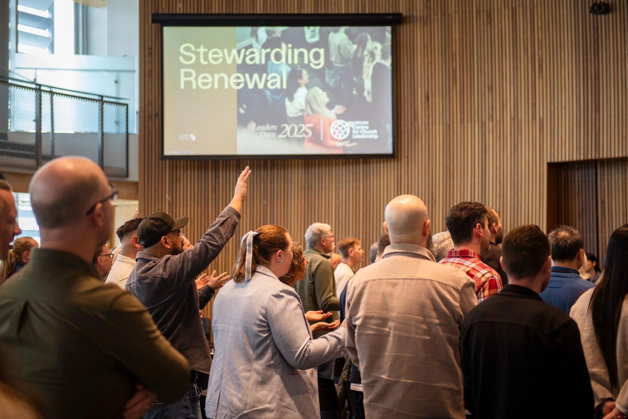 A diverse group of people attending a conference or seminar, with one person raising their hand. In the background, a large screen displays the words 'Stewarding Renewal' with an image of audience members and logos at the bottom.