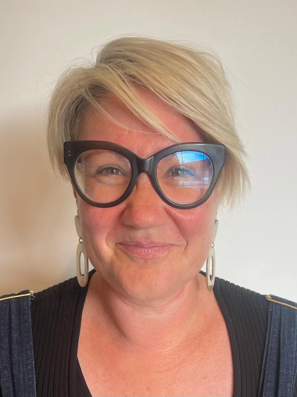 Amy, a woman with short blonde hair wearing large black glasses, earrings, and a black top, smiling against a plain white background.