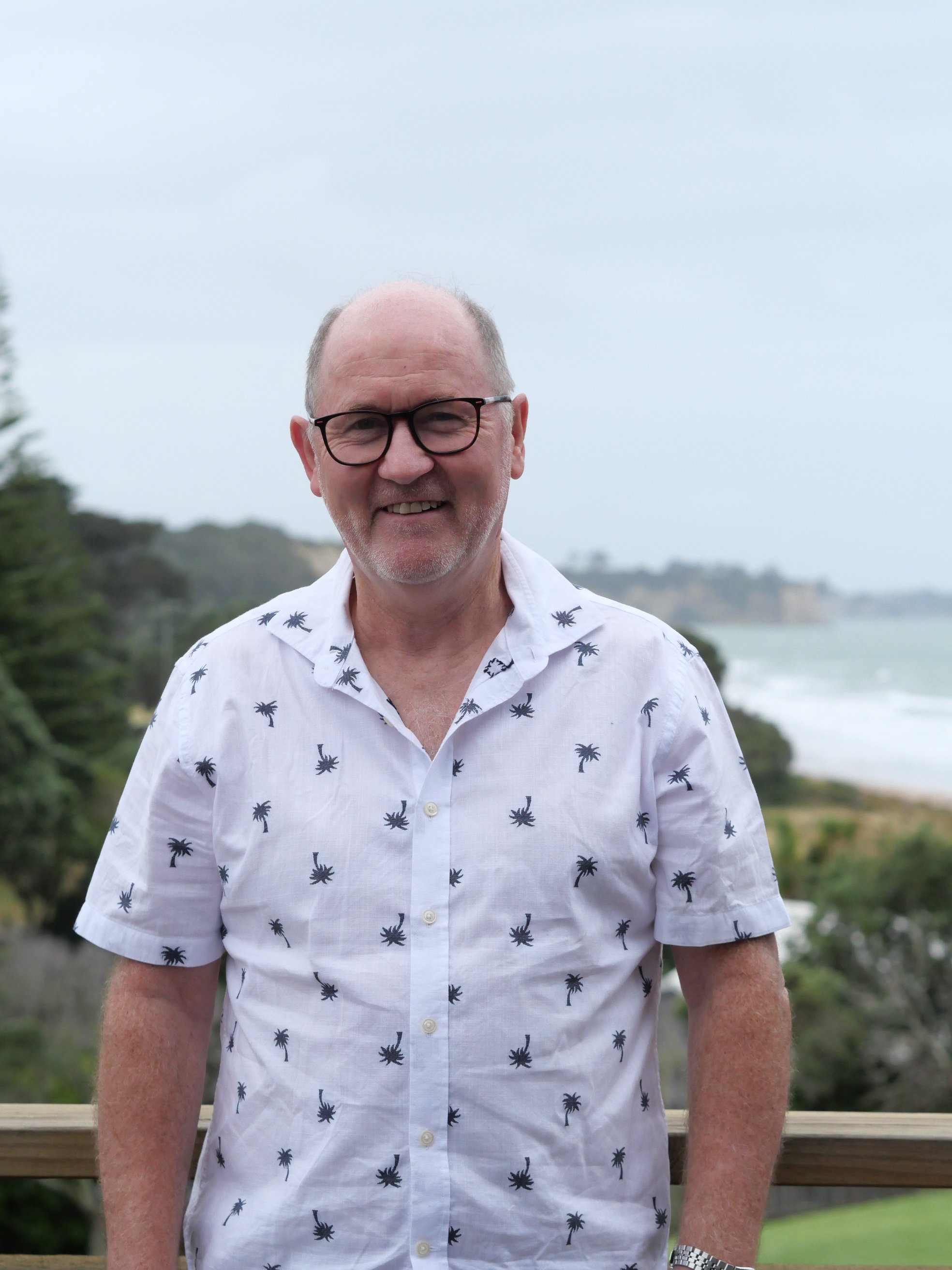 Graeme Flett, A middle-aged man wearing glasses and a white shirt with black palm tree patterns posing outdoors during daytime, with trees and water in the background.