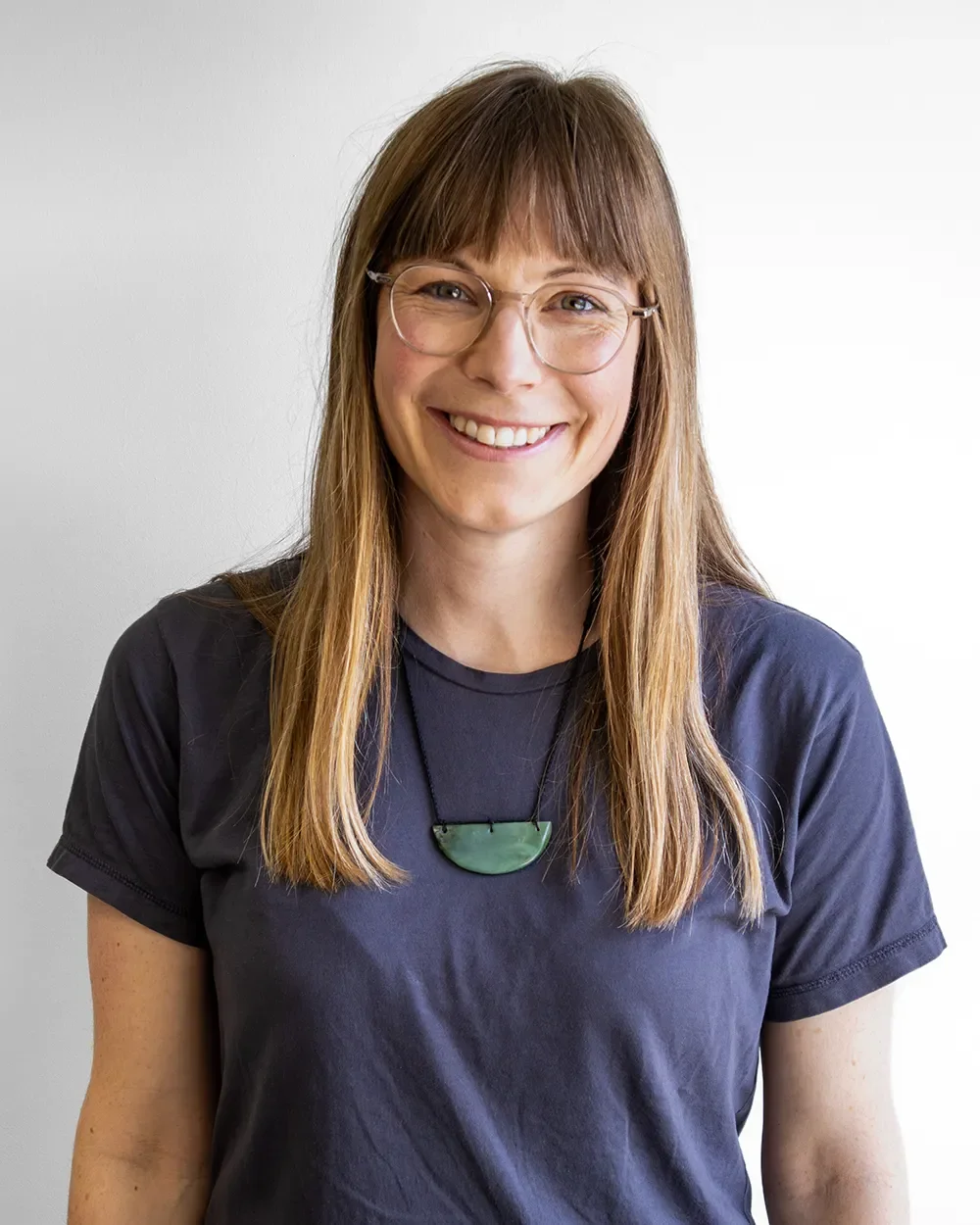 Maja Whitaker, a smiling woman with long brown hair, wearing glasses and a dark t-shirt, standing against a plain light background.