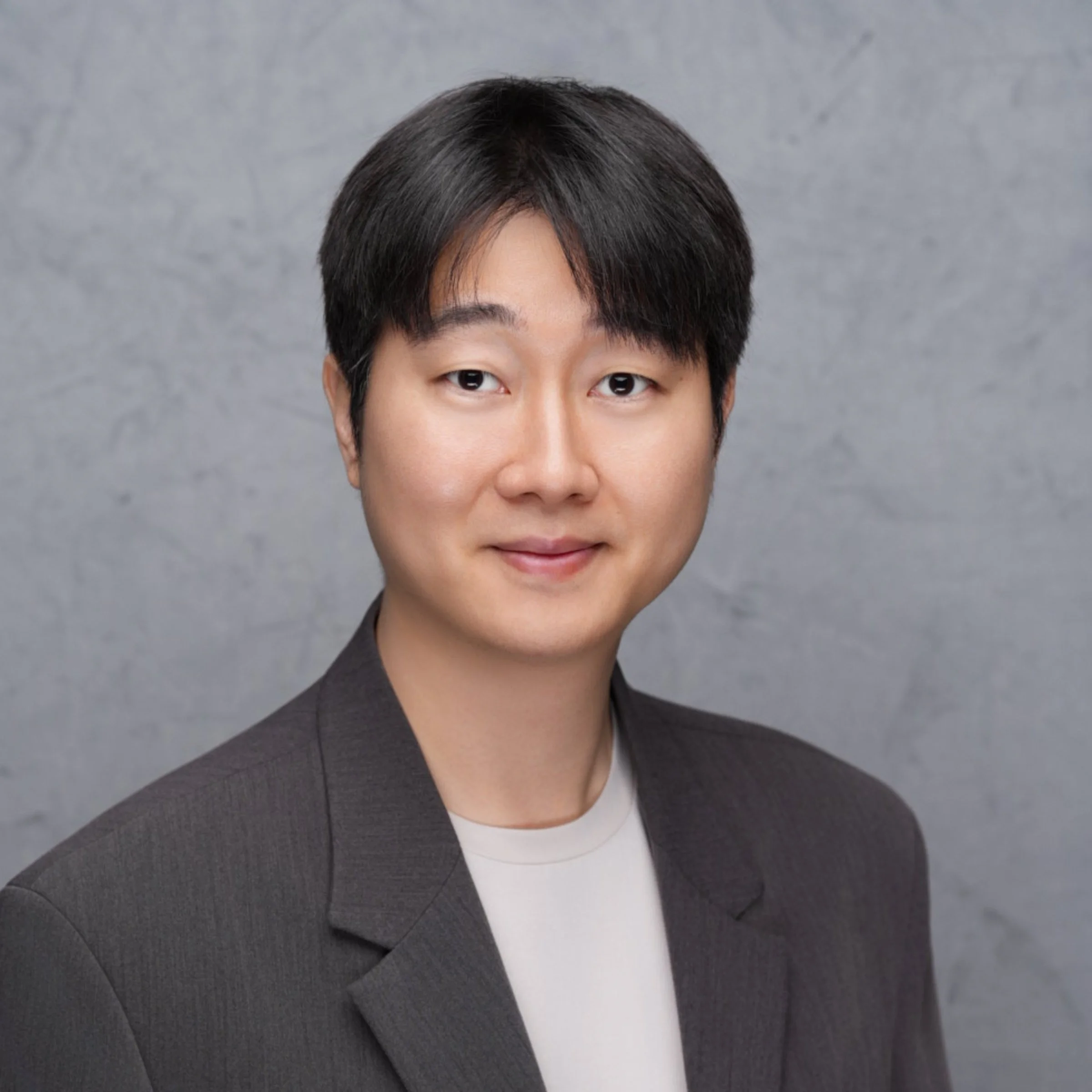 Professional headshot of a woman with short black hair, wearing a dark blazer and a white top, against a gray textured background.