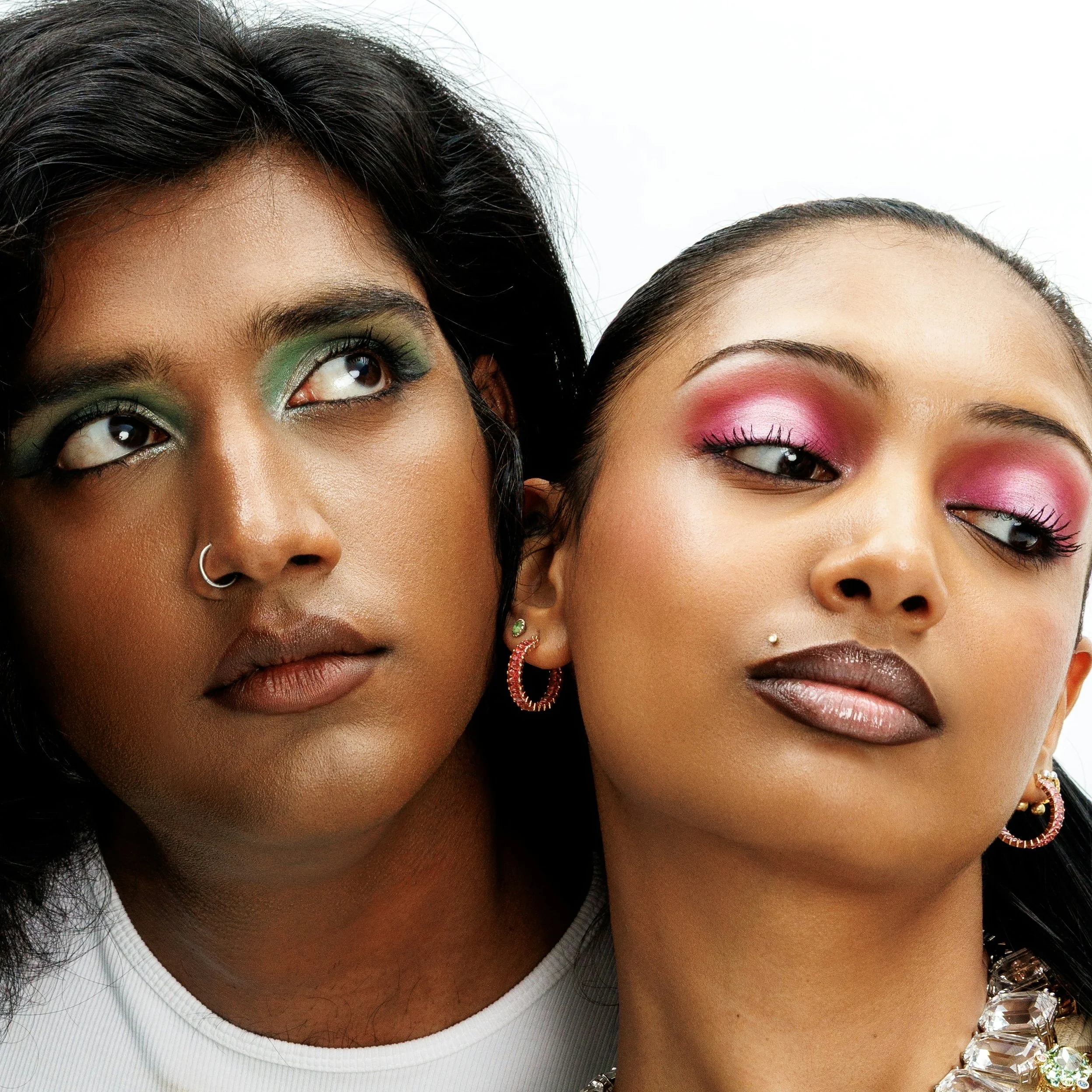 Close-up of two women with colorful eye makeup, one with green eyeshadow and the other with pink eyeshadow, wearing jewelry and facing each other