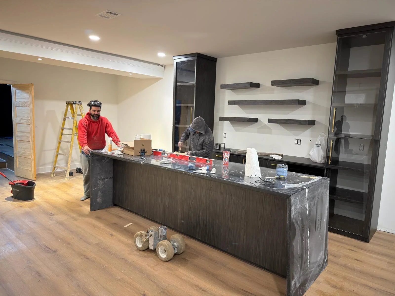Two people are working on a kitchen renovation. One is smiling and wearing a red hoodie, the other has their head down and is wearing a gray hoodie. The kitchen features dark cabinetry, open floating shelves, and a marble-like kitchen island. Tools a