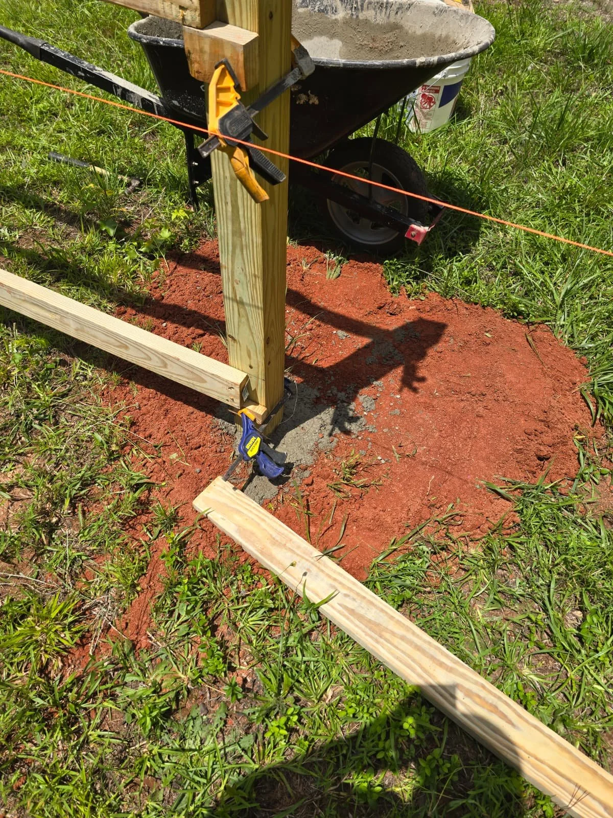 Wooden posts secured in the ground with clamps and level, with a horizontal wooden board attached, surrounded by grass, with orange string and a wheelbarrow in the background.