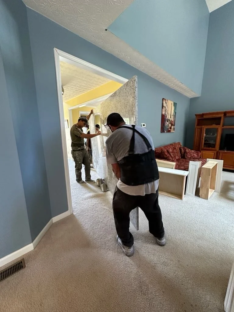Two men are moving furniture through a doorway into a living room with blue walls,  carpeted floors, a red patterned couch, and a wooden entertainment center.