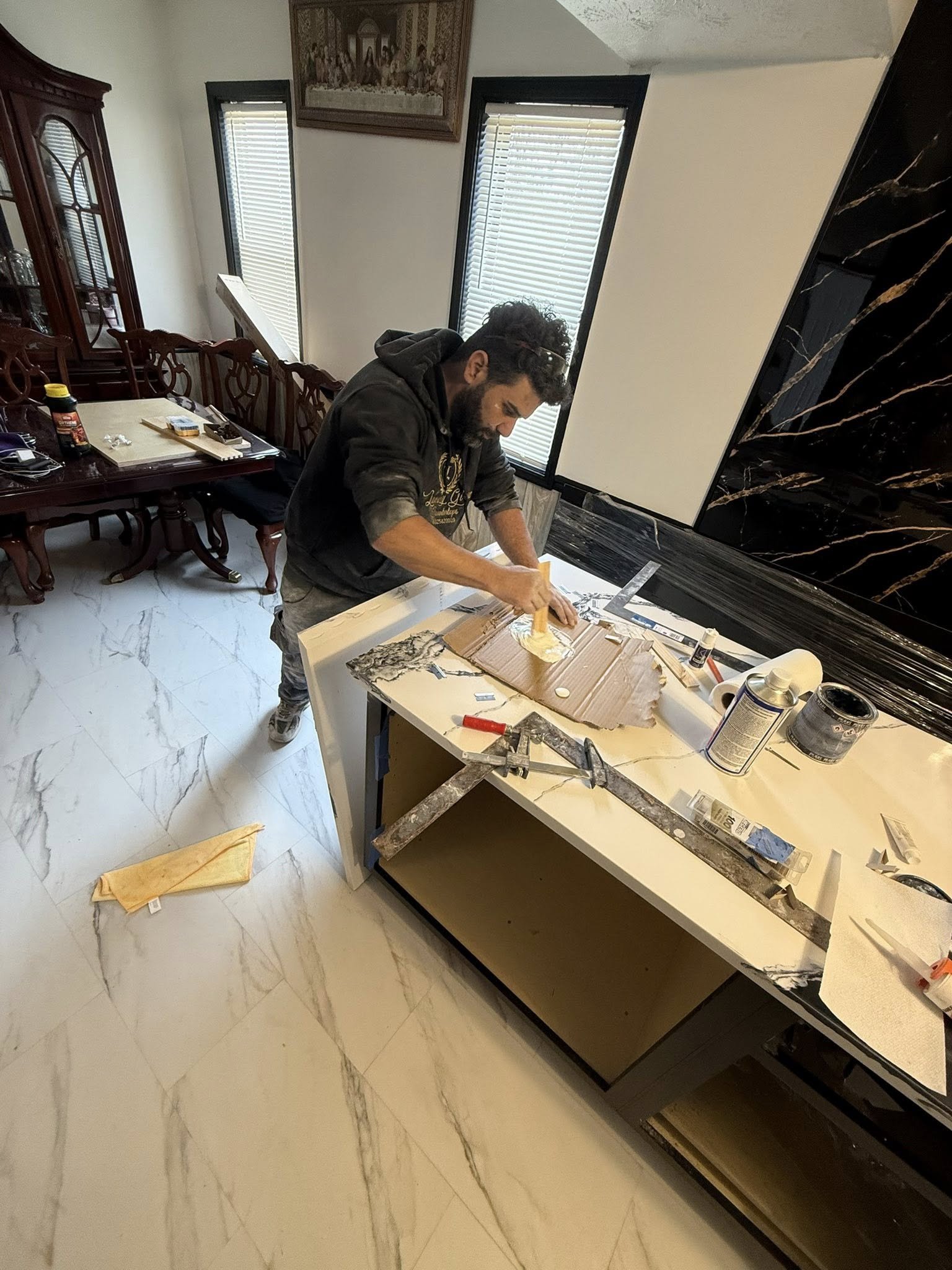A person working on a home renovation project, applying adhesive to a piece of material at a kitchen island. The kitchen has a marble floor and a black marble backsplash. There are various tools and supplies on the island.