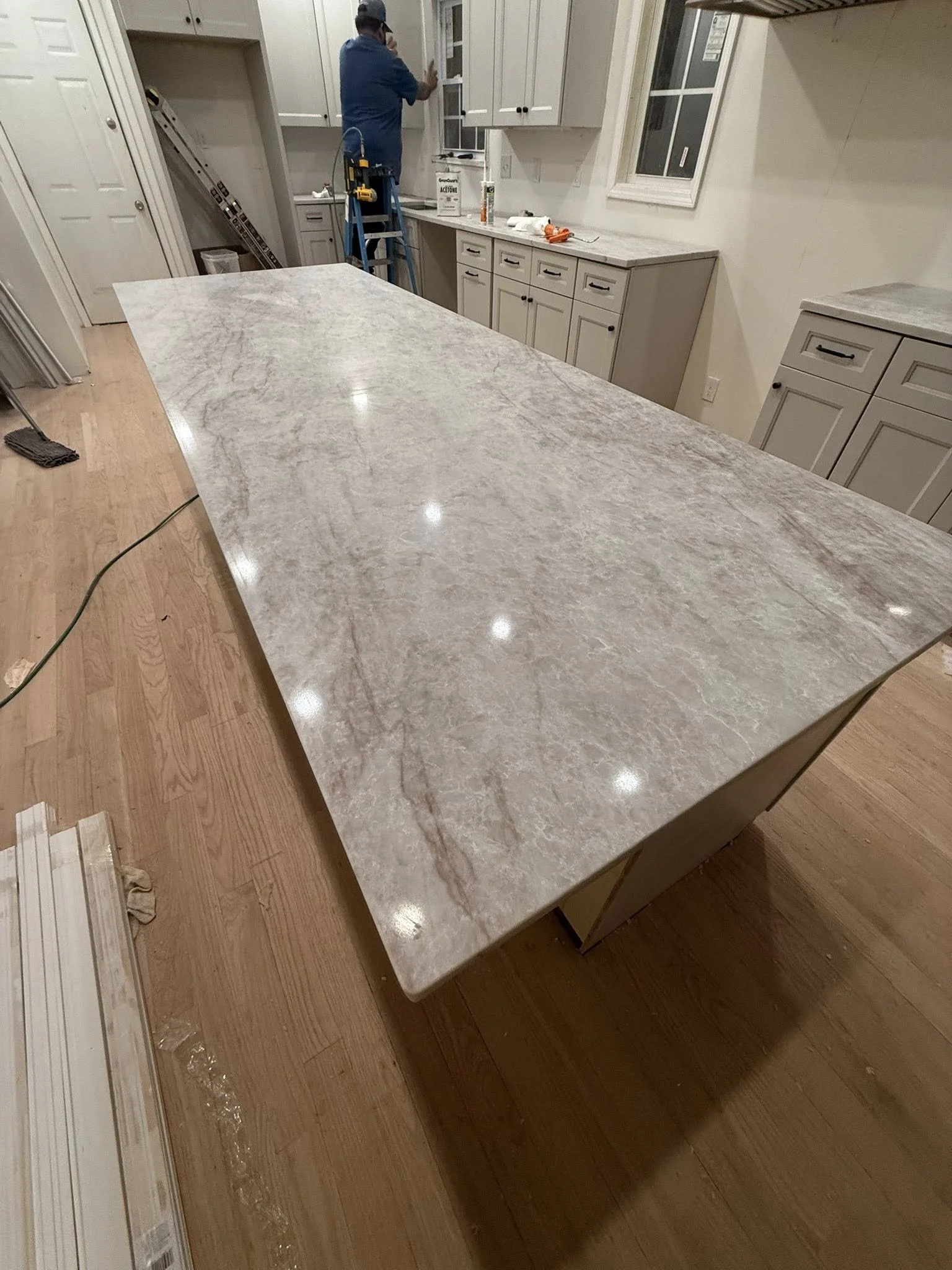 Construction worker installing or repairing a kitchen window, standing on a ladder in a modern kitchen with white cabinets and a large marble countertop.