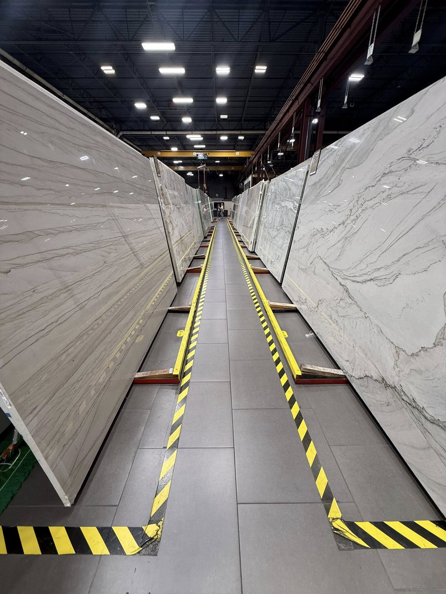 A warehouse displaying large marble slabs arranged vertically, with a walkway in the center marked by yellow and black safety tape.