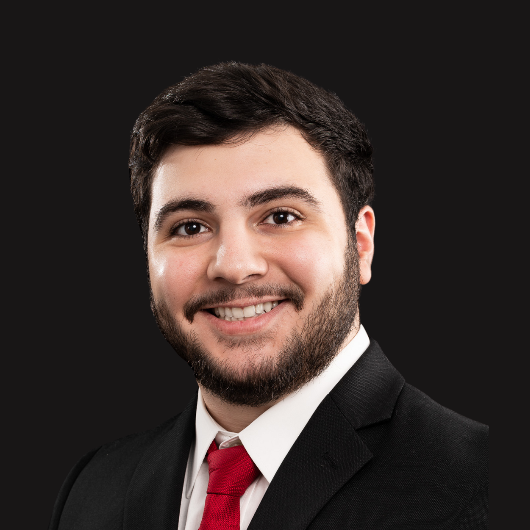 Professional portrait of a young man with dark hair and beard, wearing a black suit, white shirt, and red tie, smiling against a dark background.