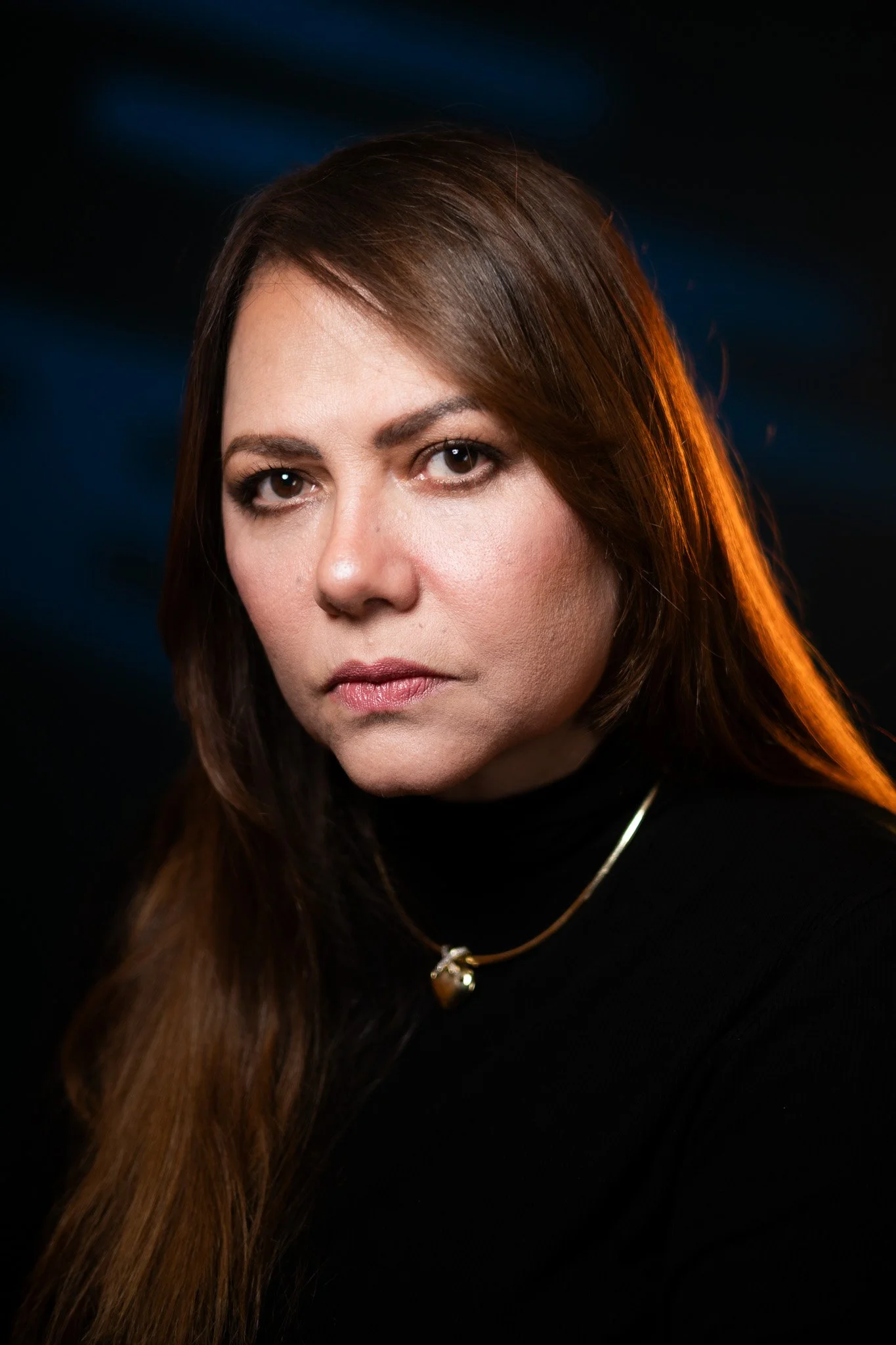 Close-up portrait of a woman with long, brown hair, wearing a black top, gold necklace, and natural makeup, against a dark background with subtle blue lighting.