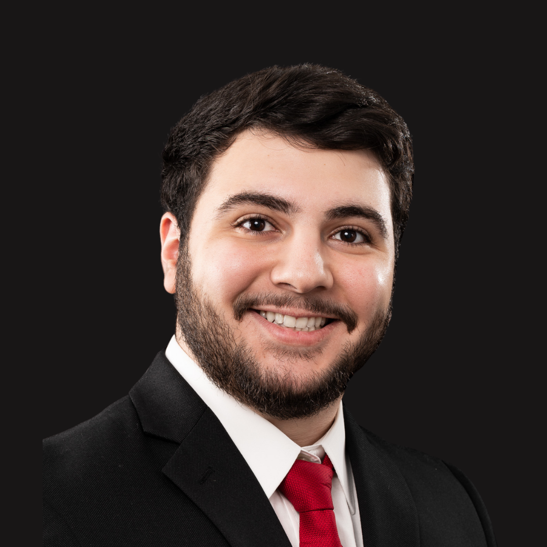 Portrait of a young man with dark hair, beard, wearing a black suit, white shirt, and red tie, smiling against a black background.