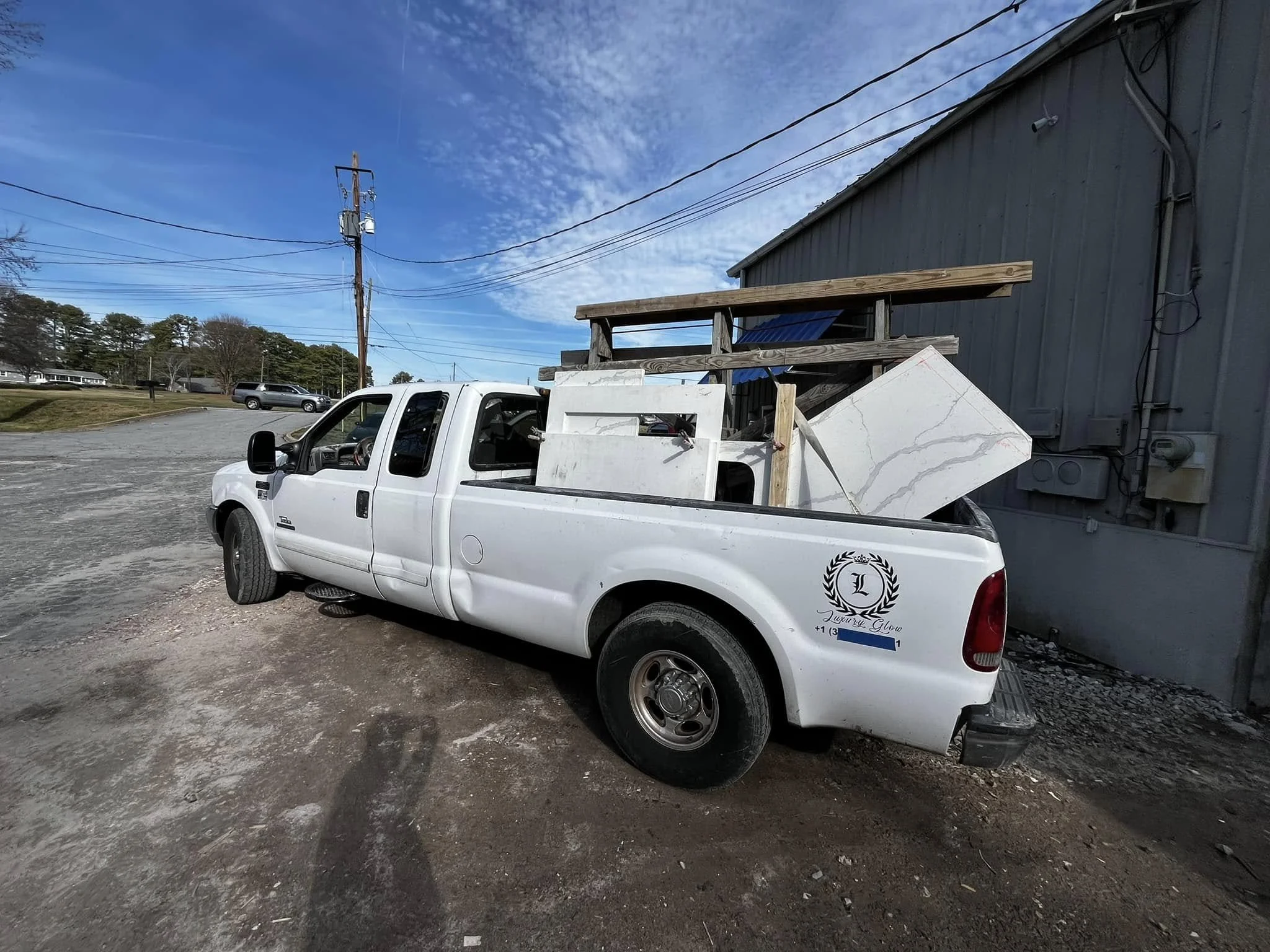 White pickup truck parked beside a gray building with various materials loaded in the truck bed, including what appears to be countertops, wood planks, and other construction supplies.
