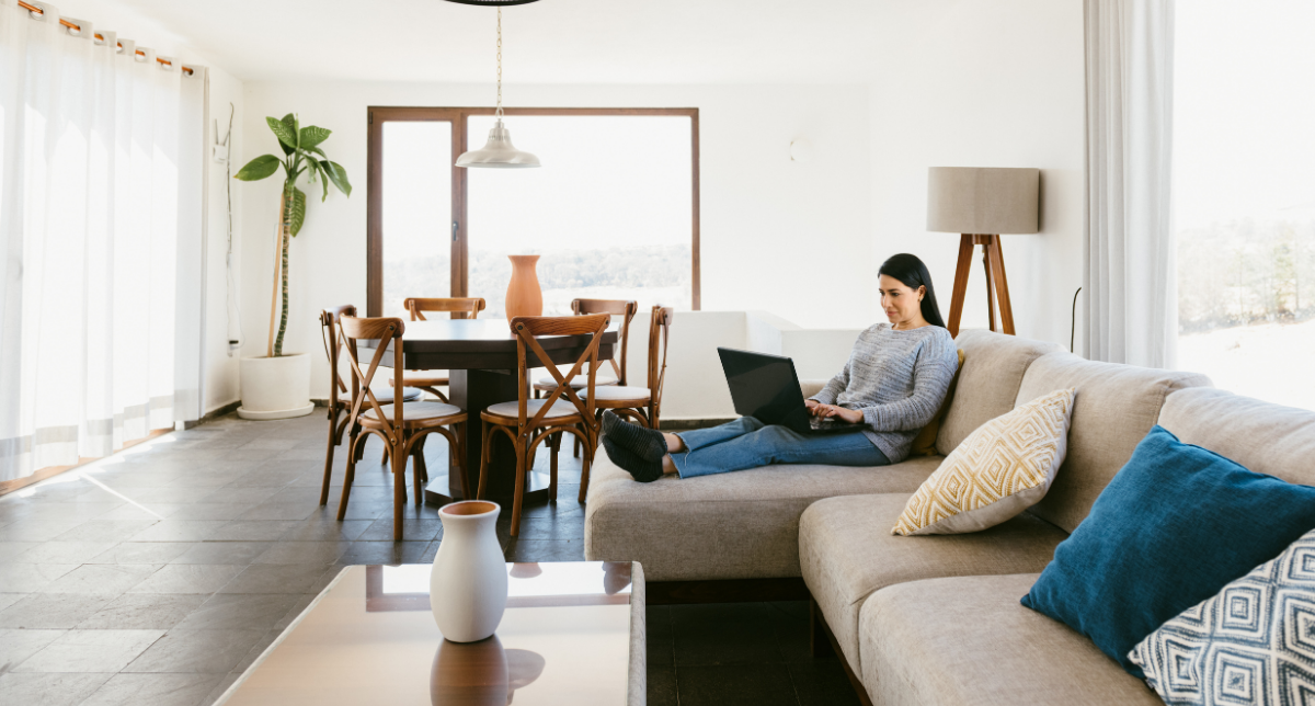 Woman sitting on a beige couch with patterned pillows, working on a laptop, in a bright living room with large windows, a wooden dining table with chairs, and indoor plants.