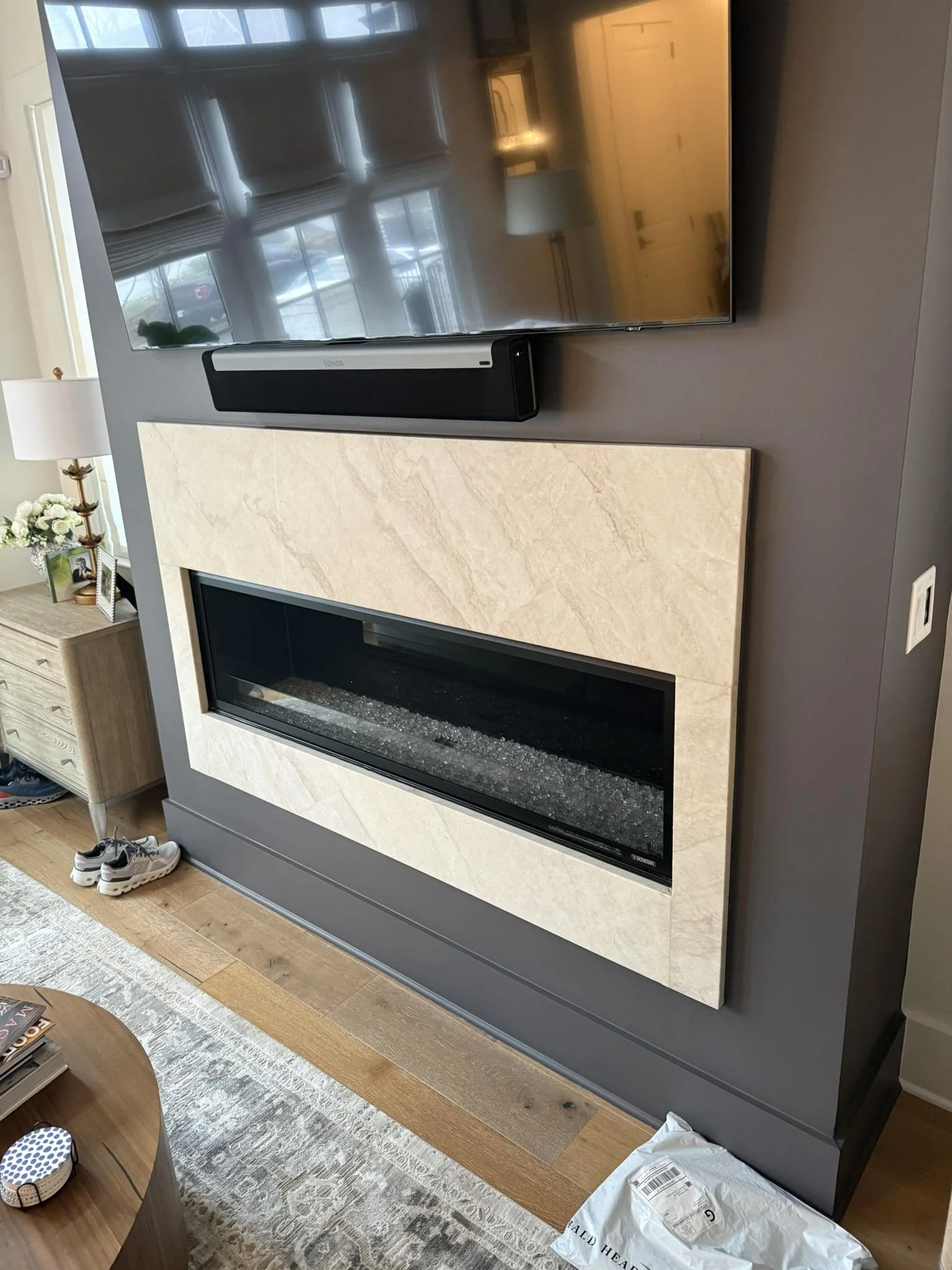 A modern living room features a wall-mounted television above a built-in fireplace with a marble surround and a black glass interior. To the left, there is a wooden side table with a flower arrangement, framed photographs, and a table lamp. Shoes are