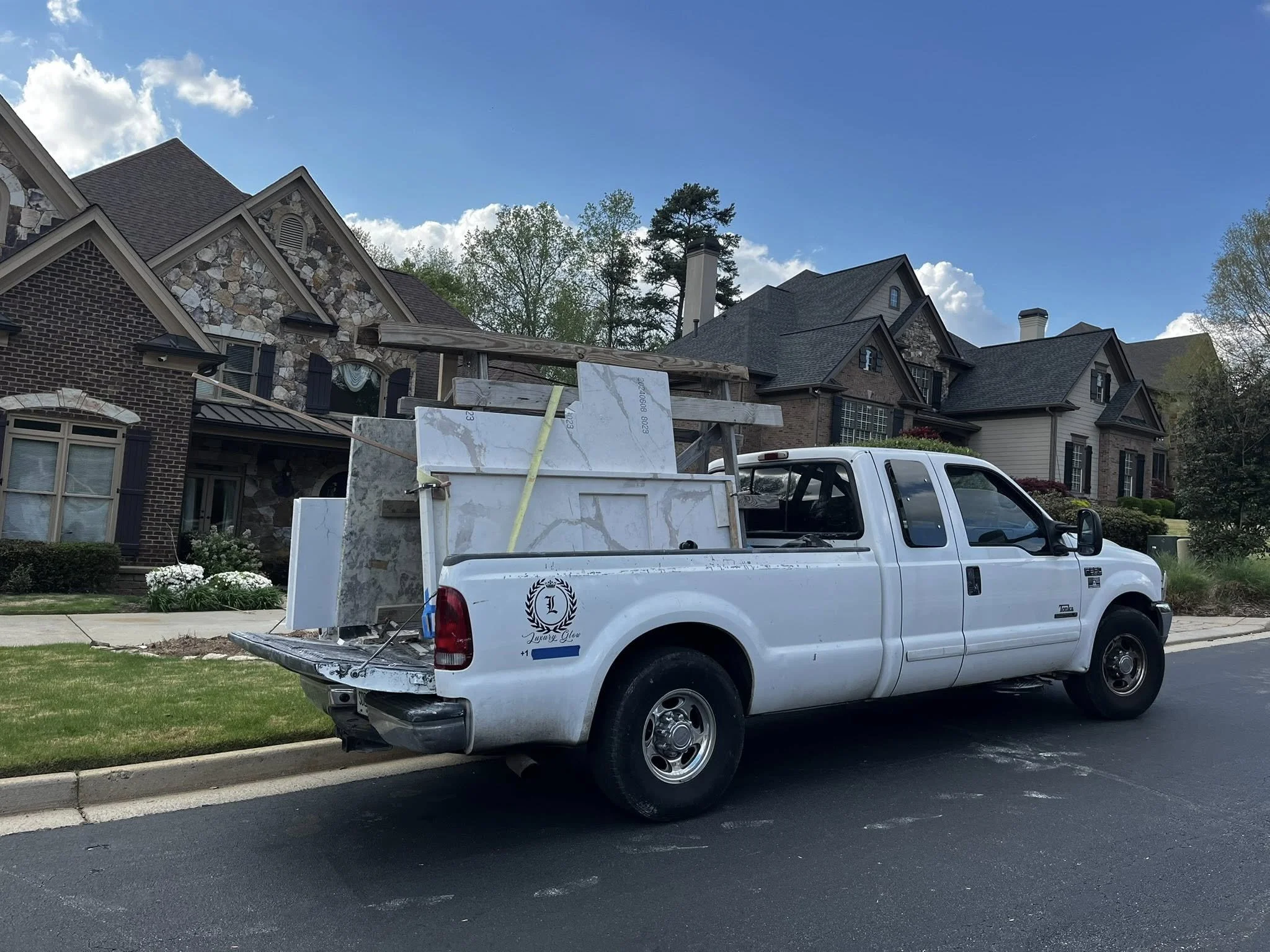 A white pickup truck parked on the side of a residential street carrying large marble or granite slabs in its bed, with a suburban neighborhood of large brick and stone houses in the background under a blue sky with some clouds.