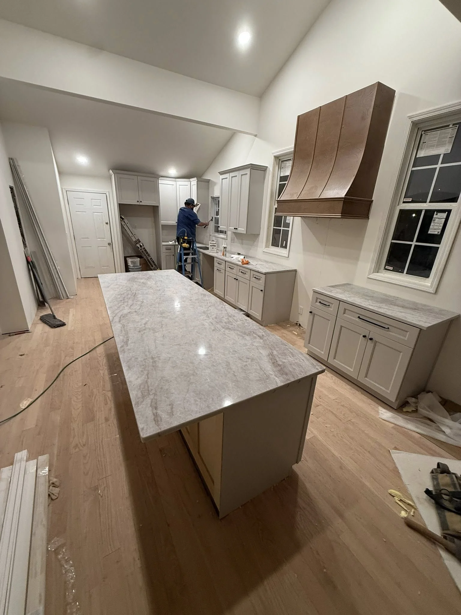 A kitchen under construction with a worker installing cabinets. There is a large central island with a marble countertop, and additional cabinets on the wall with a range hood above. The room has hardwood floors and multiple windows.