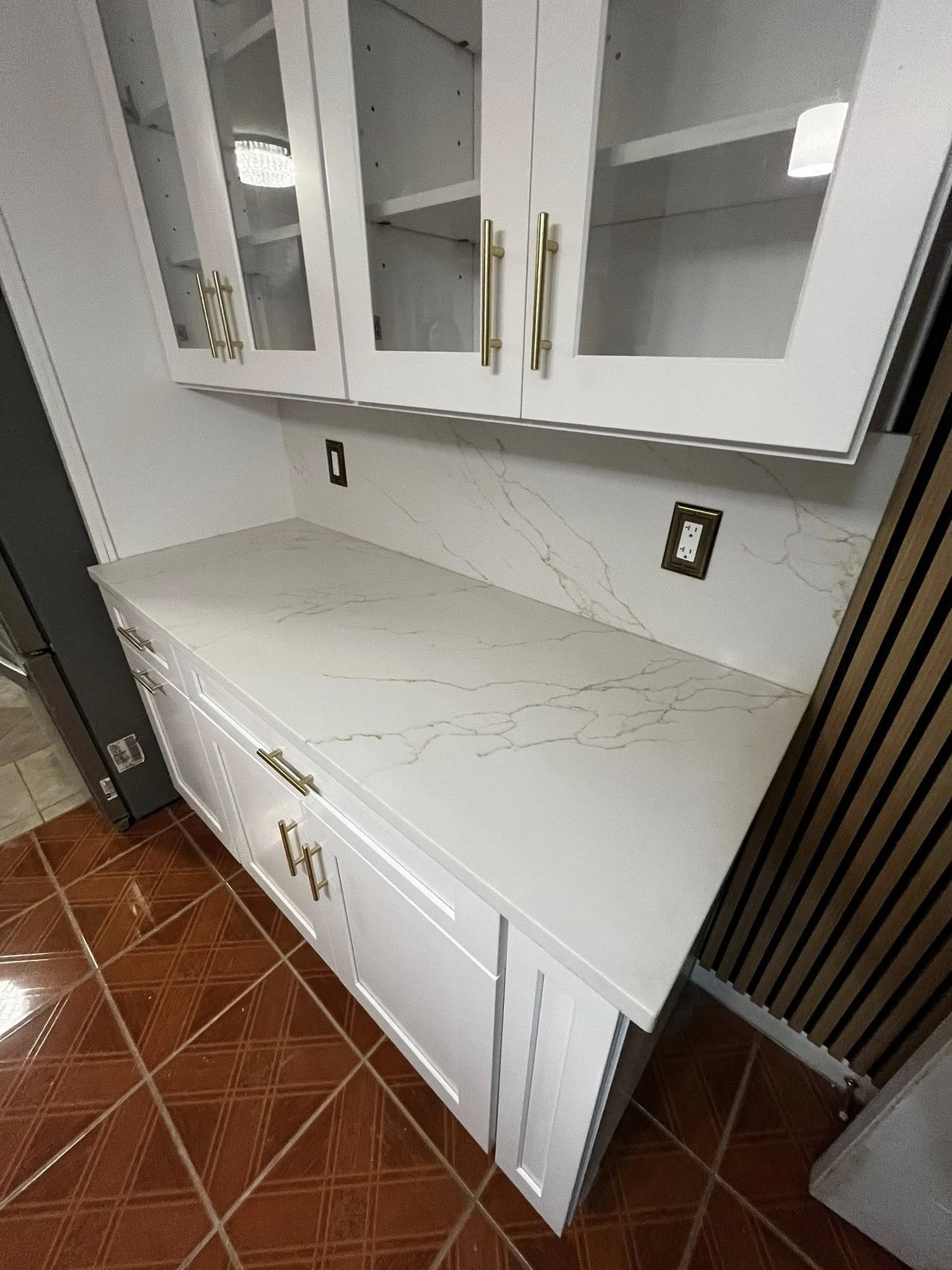 White kitchen cabinet with glass-front upper doors and gold handles, a marble countertop, and two electrical outlets on the wall. The floor has reddish-brown tiles.