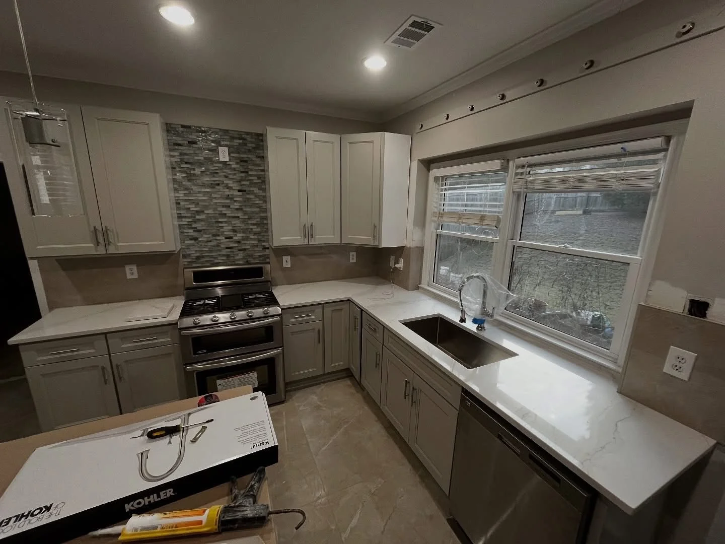 Kitchen with white cabinets, stainless steel stove, marble countertop, window over sink, tile backsplash, and construction tools on an island.