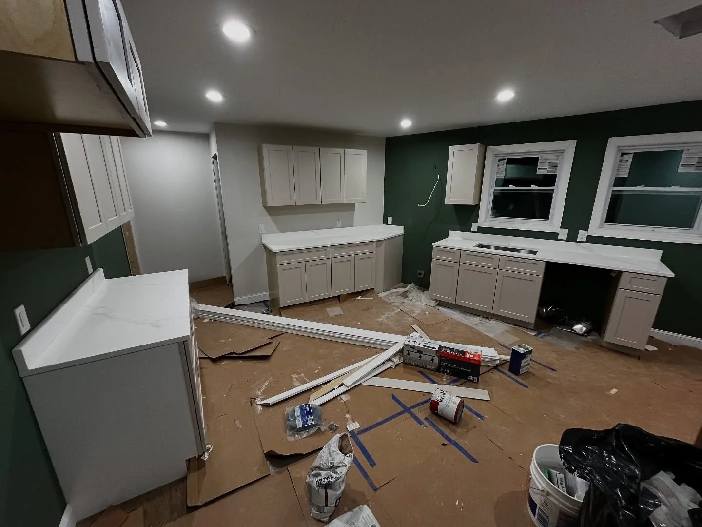 Kitchen under renovation with white cabinets and countertops, green accent wall, window frames, construction tools, materials, and debris on the floor.