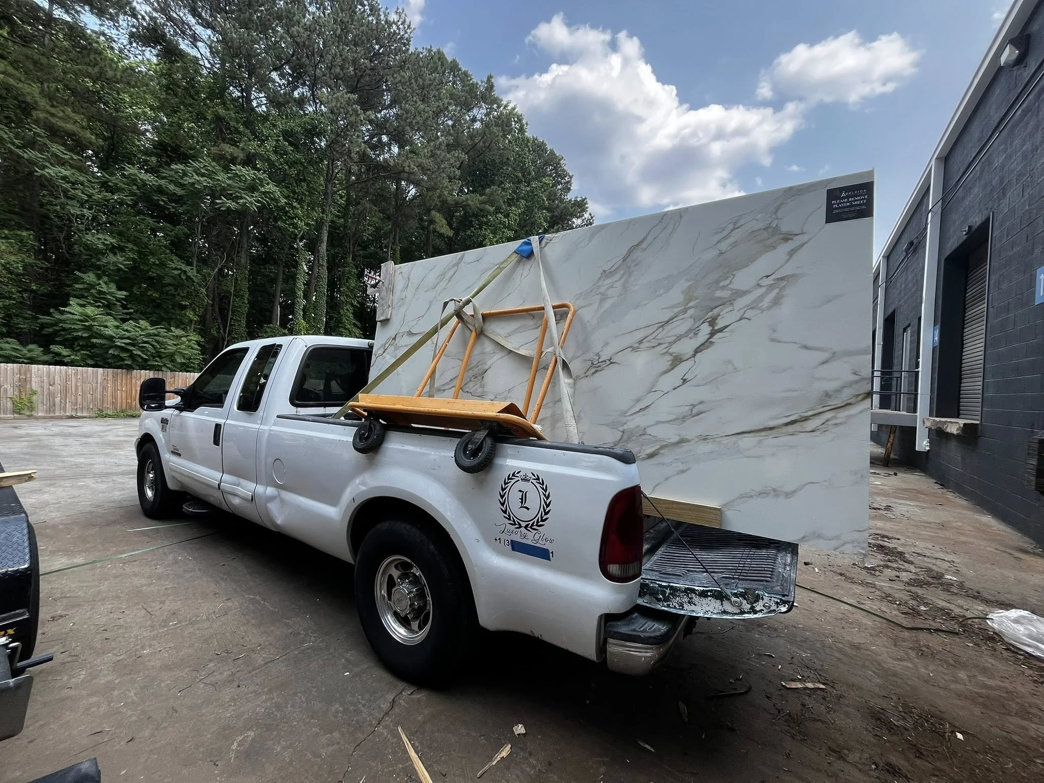 White pickup truck parked outdoors with a large marble slab in the truck bed, a cart, and a wooden board on top. Trees and a building with a rolled-down window are in the background.