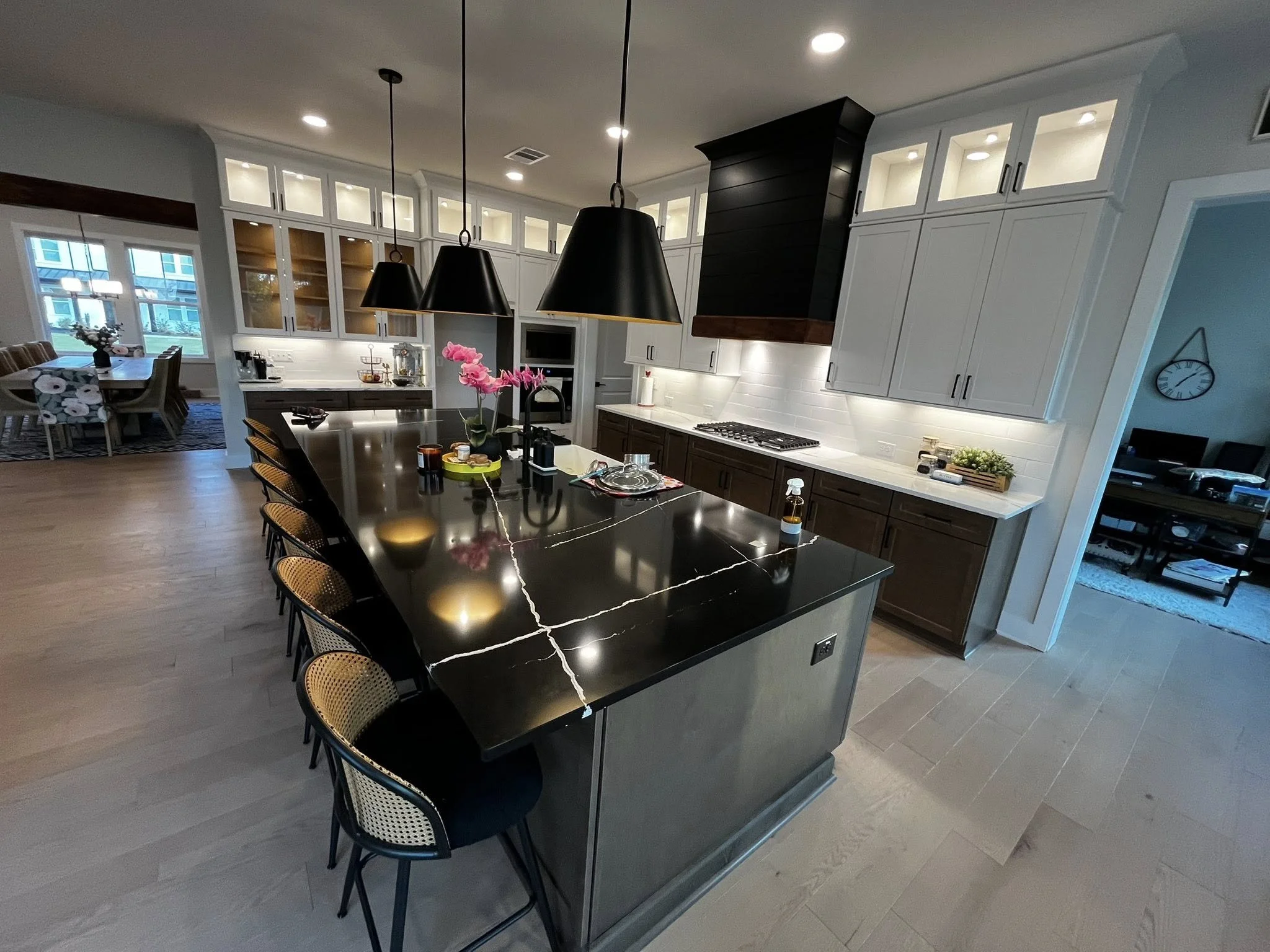 Modern kitchen with black marble island, white and dark wood cabinets, black pendant lights, and a floral centerpiece, with open view to dining and living areas.