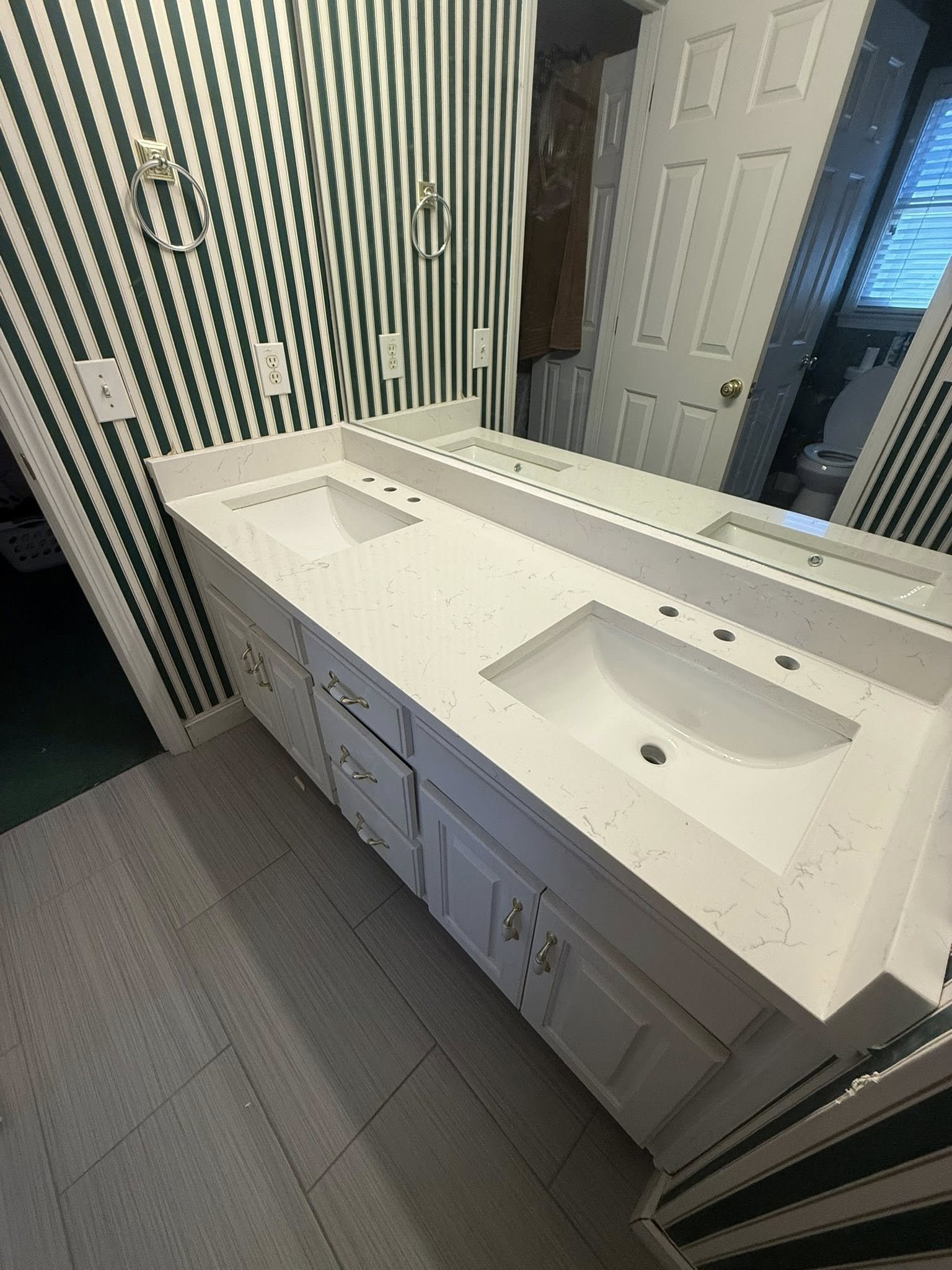 Bathroom vanity with a double sink and a large mirror, striped wall, and a toilet visible in the background.