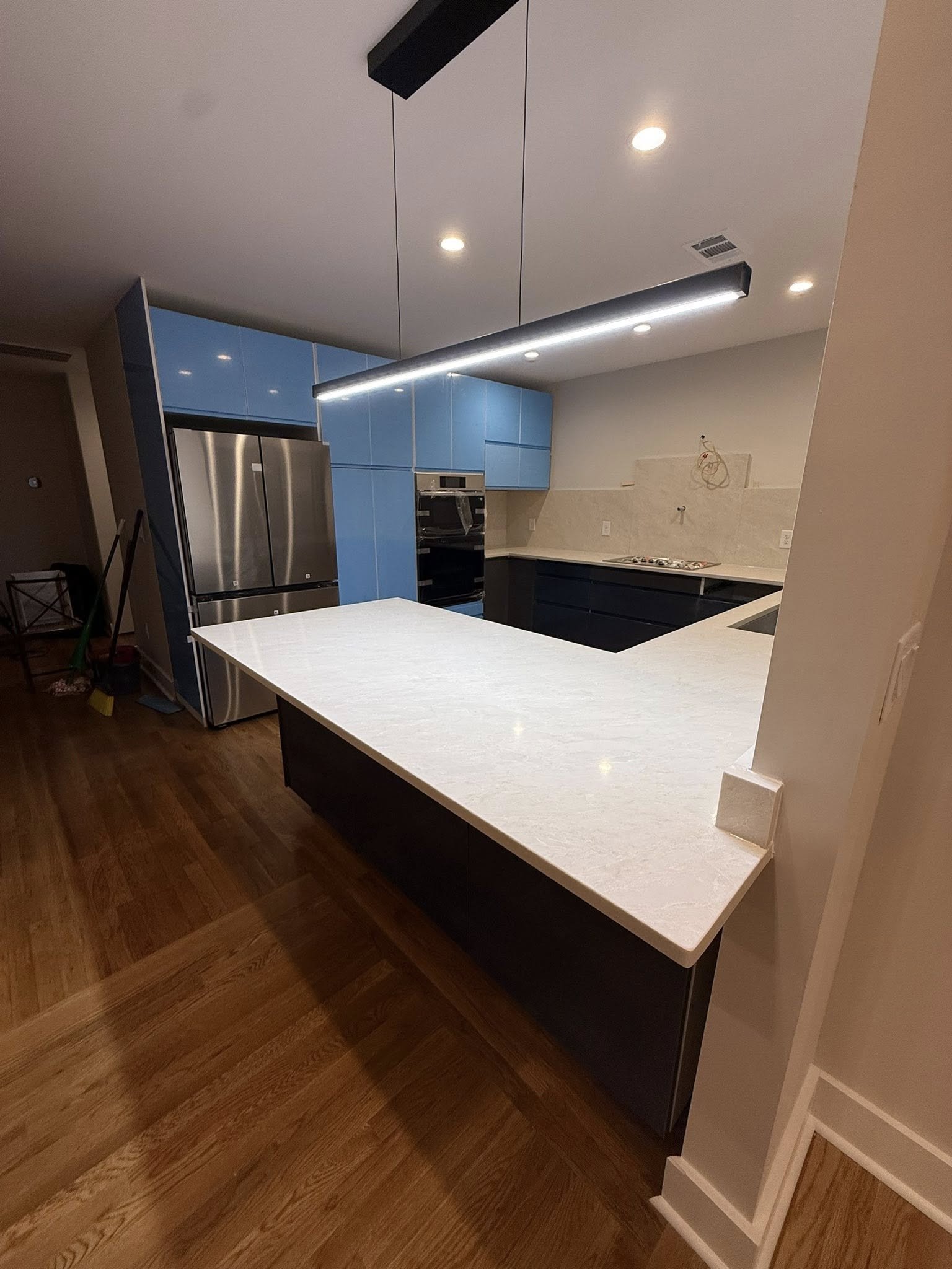 Modern kitchen with blue and black cabinetry, stainless steel refrigerator, built-in oven, and white marble countertop island, with wooden flooring and recessed lighting.