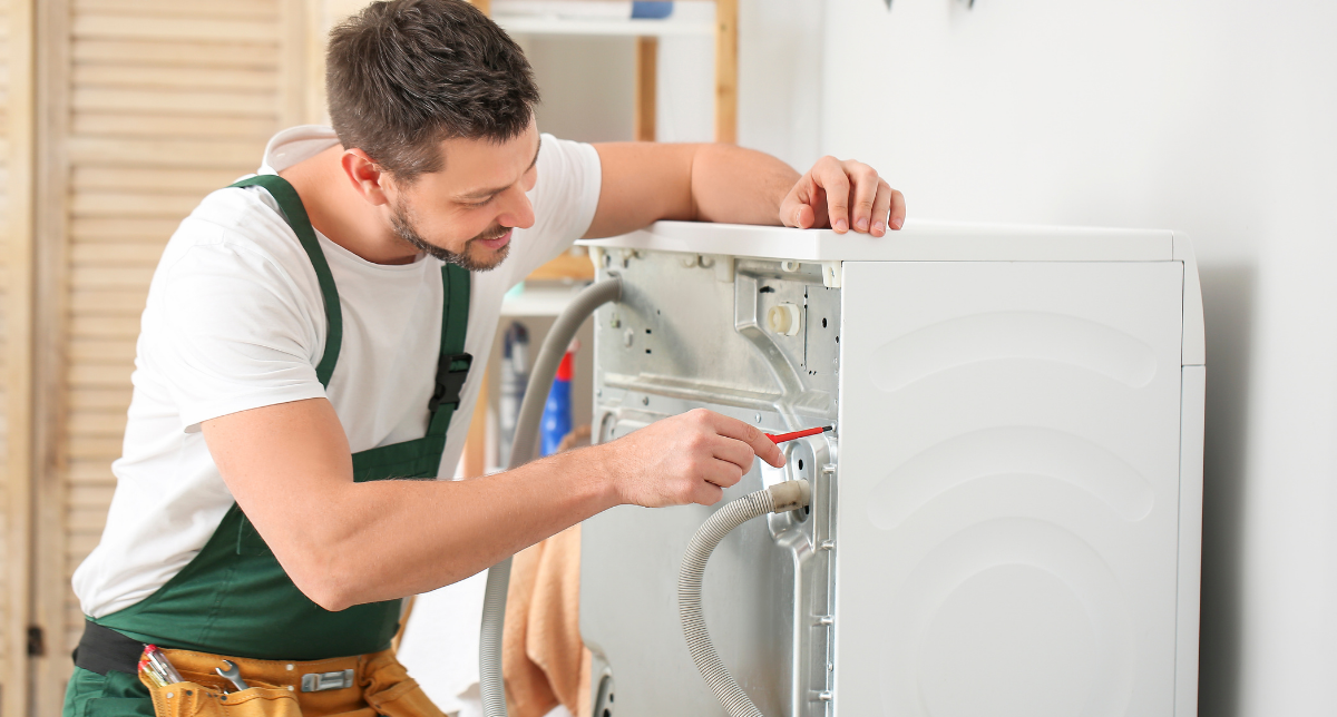 A man in a white T-shirt and green apron repairs a washing machine using a screwdriver.
