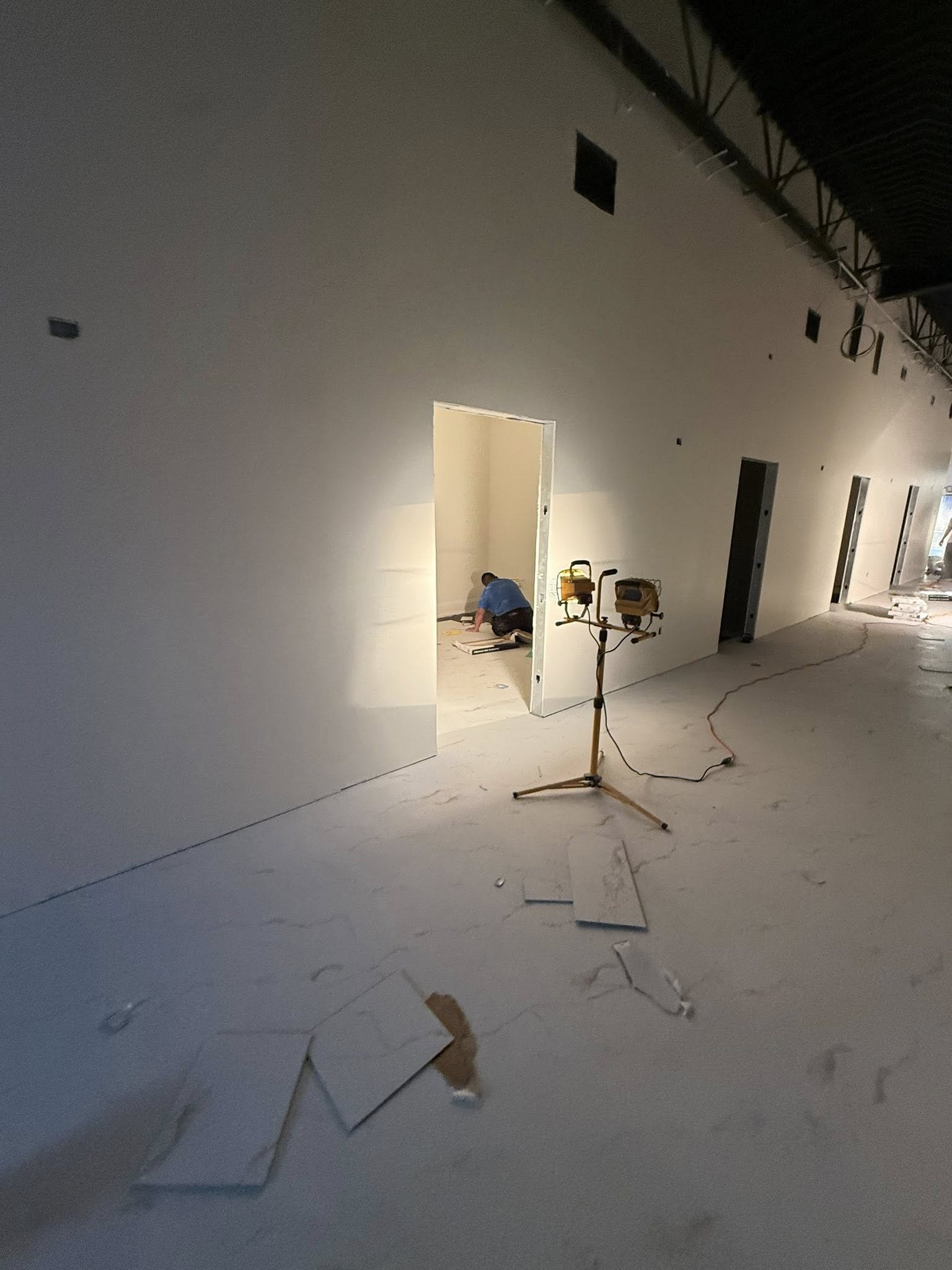 Construction worker installing drywall in a building under construction, with drywall panels and construction tools visible on the floor.