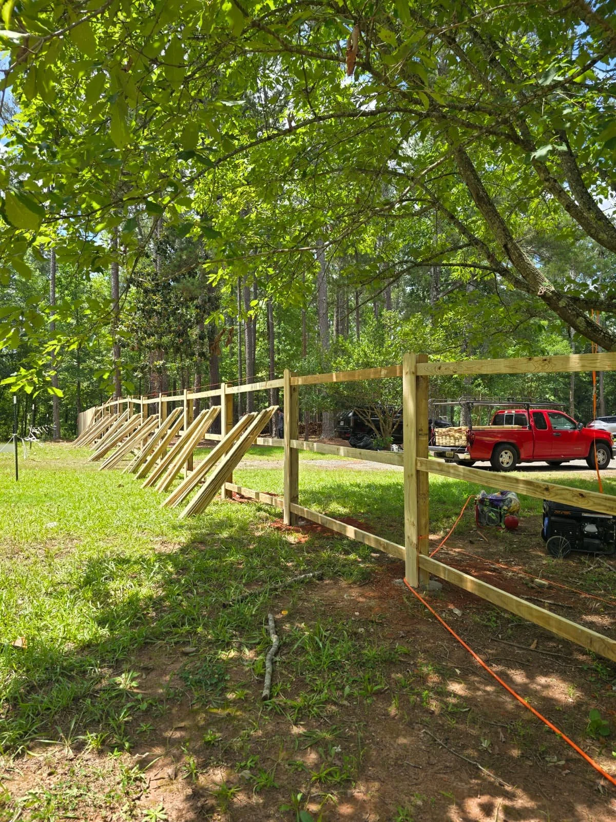 A wooden fence under construction in a grassy yard with trees, a red pickup truck, and construction tools and equipment nearby.