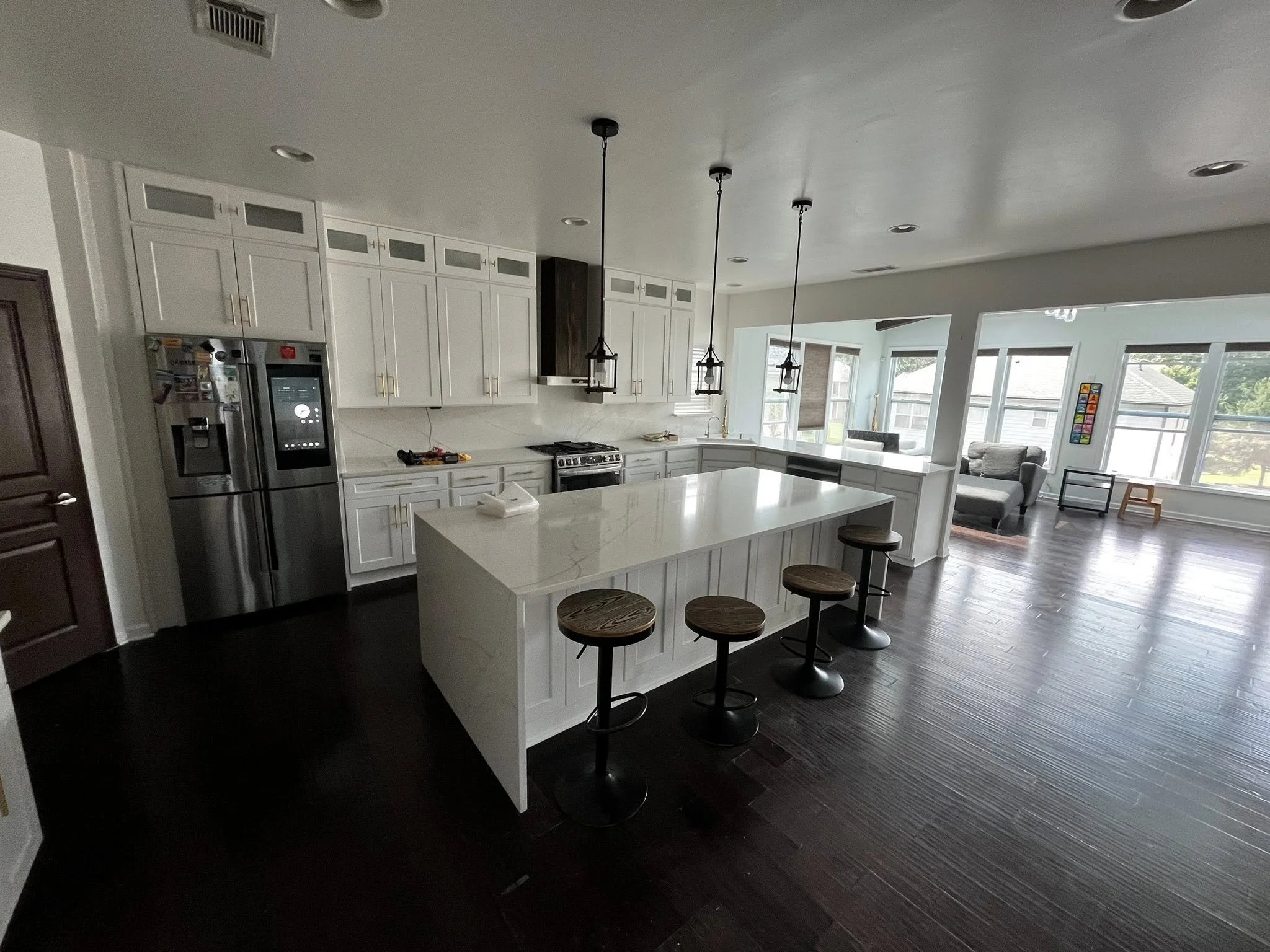 Open-concept kitchen with white cabinets, a large kitchen island with a white countertop, pendant lights, and a modern stainless steel fridge. In the background, a bright living area with multiple windows, seating, and a dark wood floor.