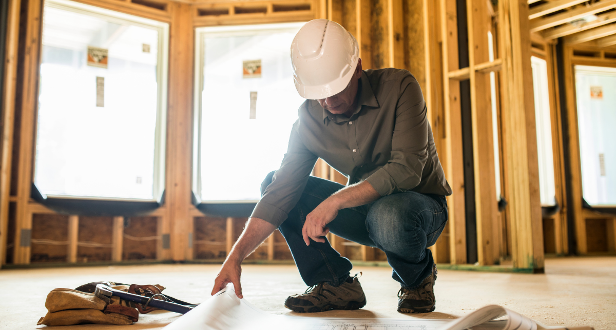 A man wearing a white construction helmet and casual clothes kneeling inside a house under construction, examining blueprints on the floor.