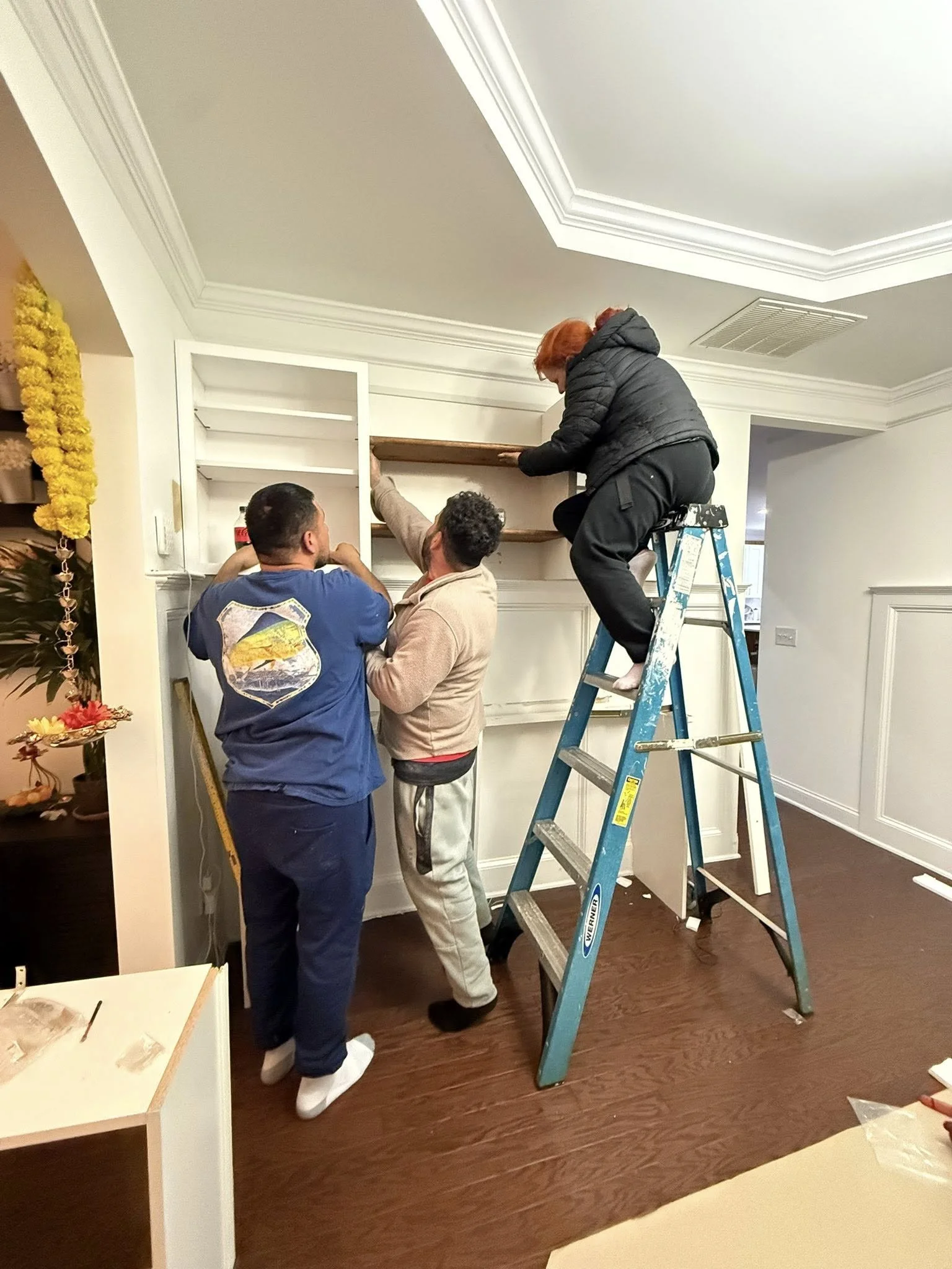 Three people installing wooden shelves on a wall. One woman is on a step ladder securing a shelf, while two men are below assisting and holding the shelf in place.