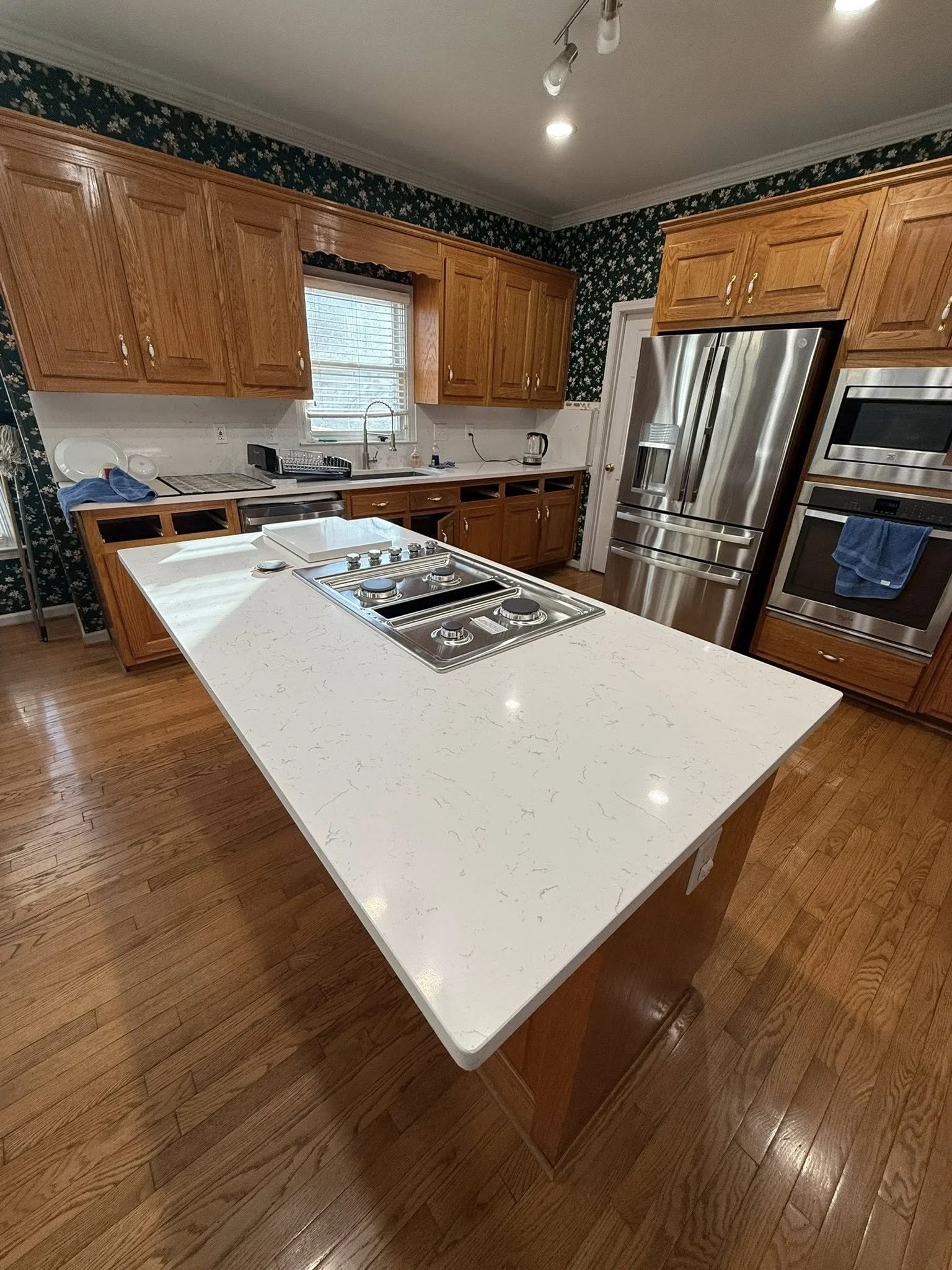 A kitchen with wooden cabinets, a white countertop island with a stove, stainless steel refrigerator, built-in oven, and microwave, dark floral wallpaper, and hardwood flooring.