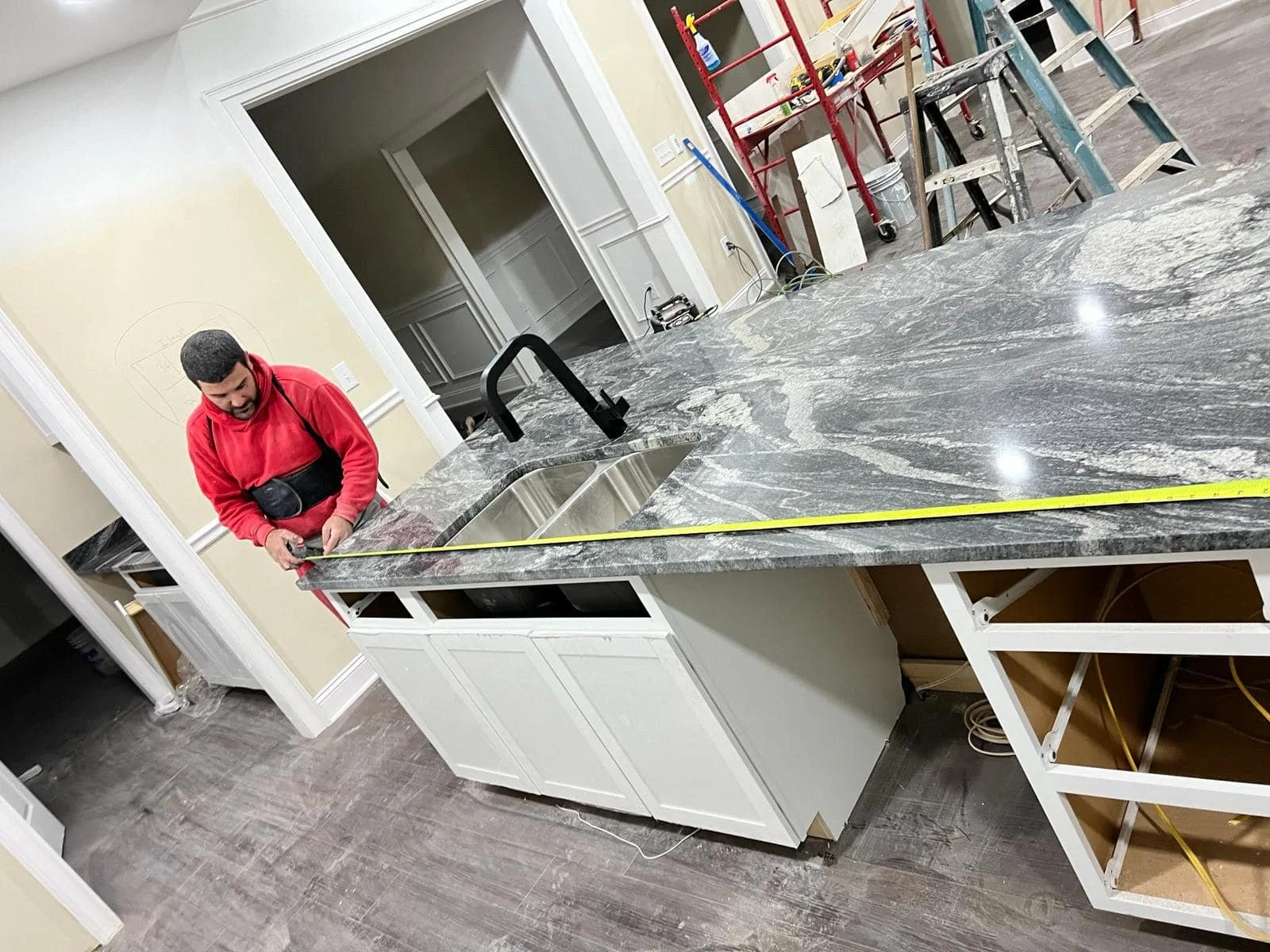 Construction worker measuring a marble kitchen island countertop in a home renovation. There are ladders and tools in the background, with open cabinetry underneath the counter and a sink with a black faucet.
