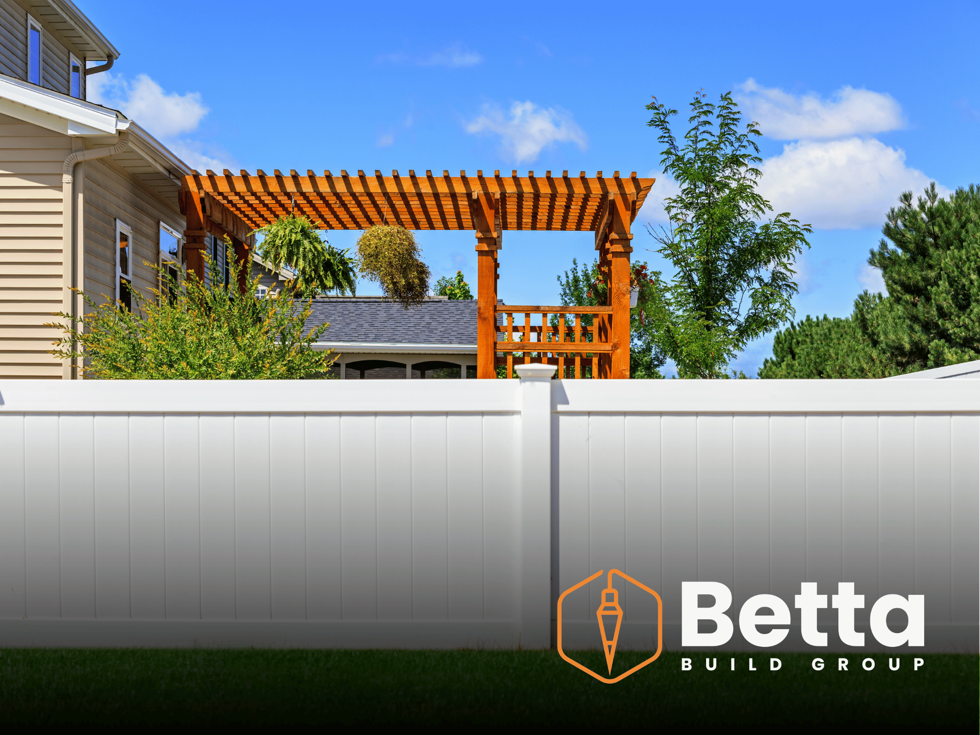 A backyard with a white fence, a wooden pergola, several green trees, and a house with beige siding and white trim under a partly cloudy blue sky.