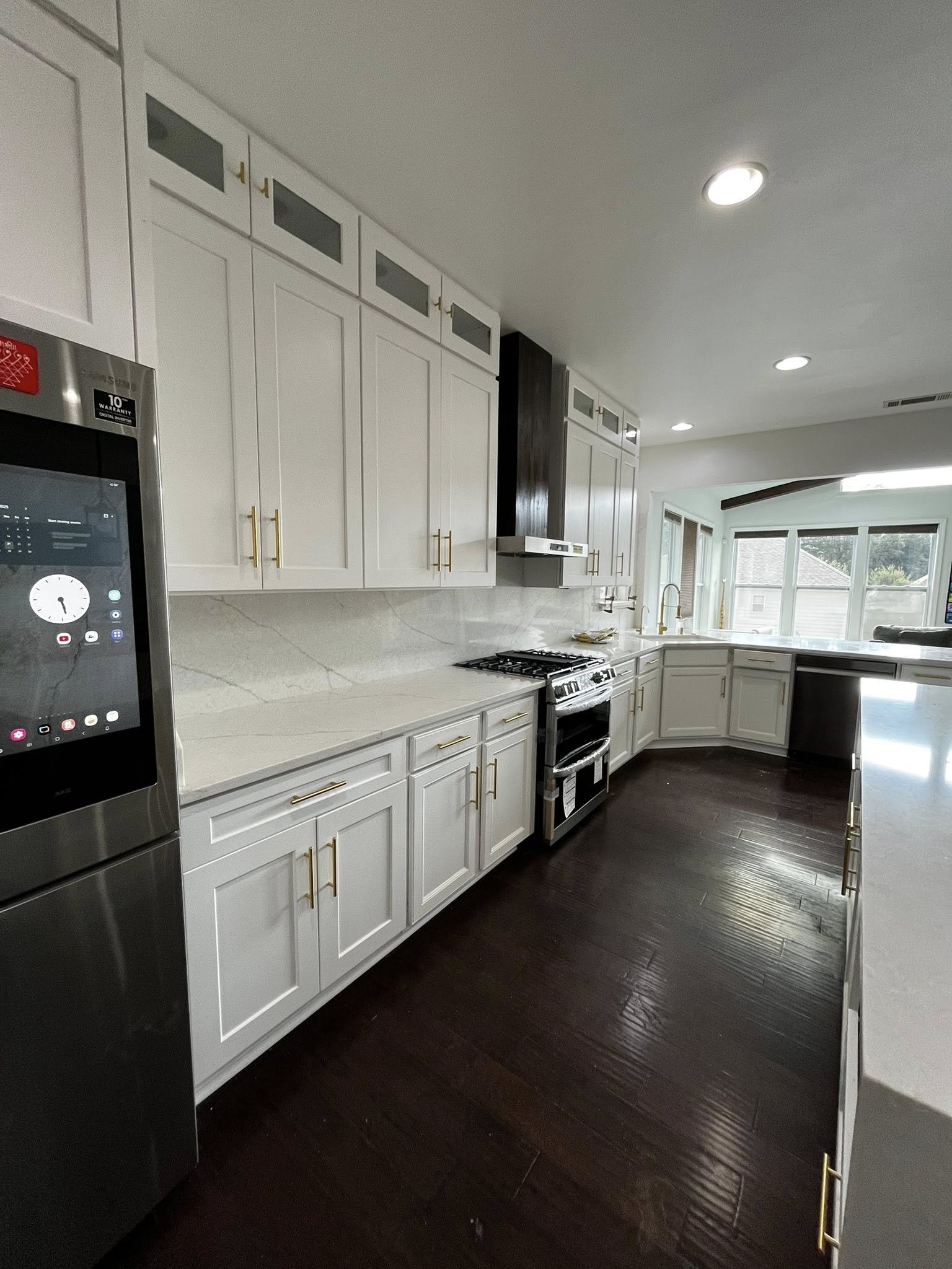 A modern kitchen with white cabinets, dark wood flooring, a stainless steel refrigerator, a stove, and large windows allowing natural light.