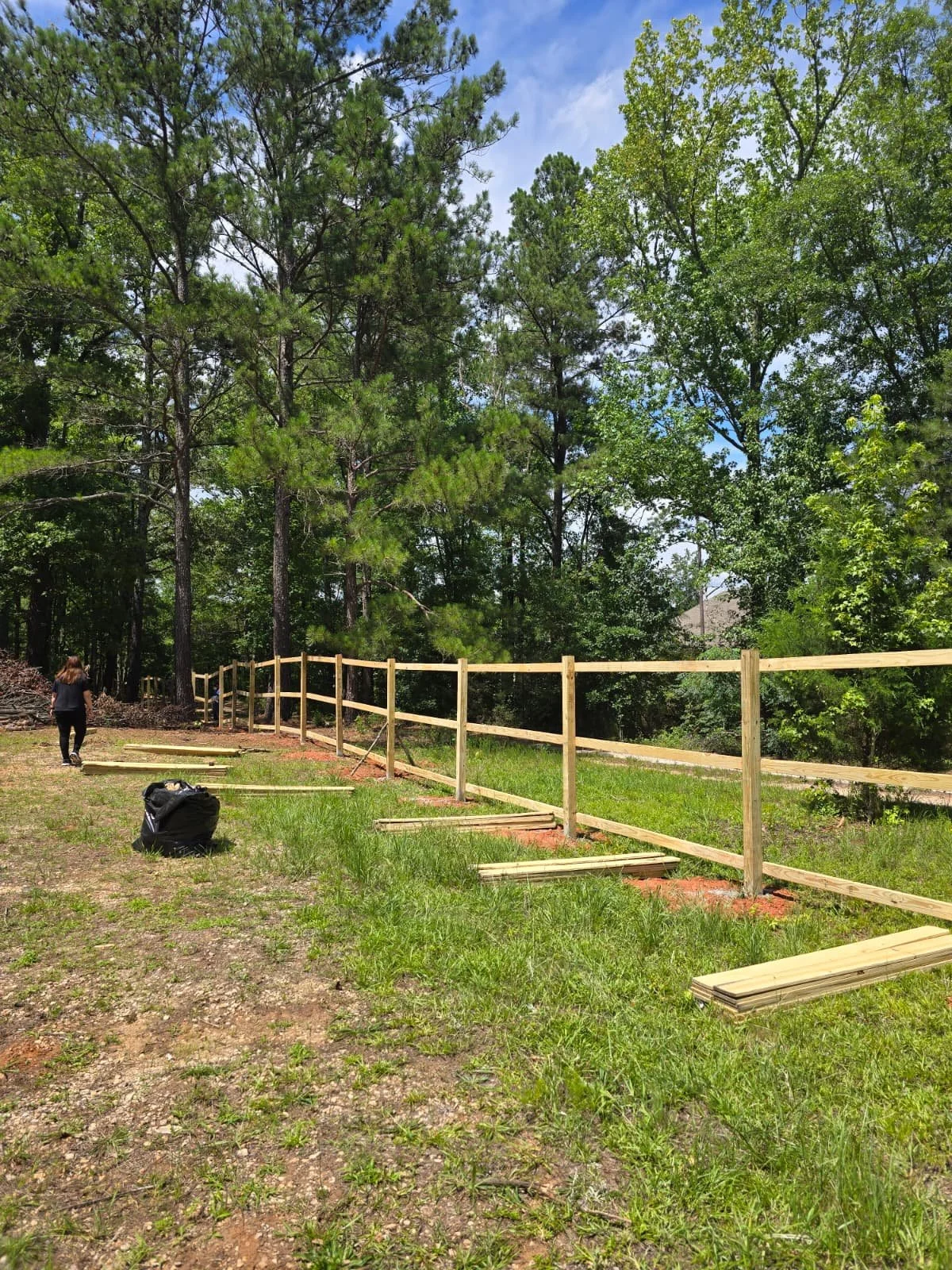 A woman building a wooden fence along a grassy area with trees in the background, under a partly cloudy sky.