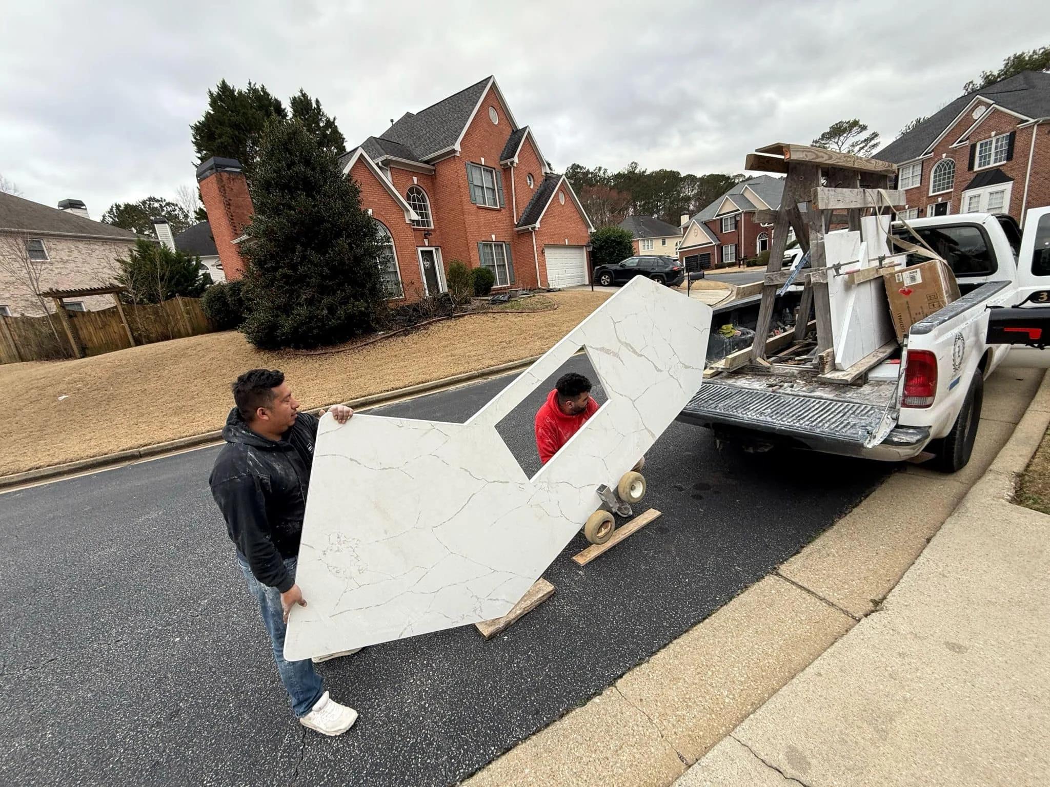 Two men are unloading a large marble or stone piece from the back of a white pickup truck onto the asphalt street in a residential neighborhood with brick houses.